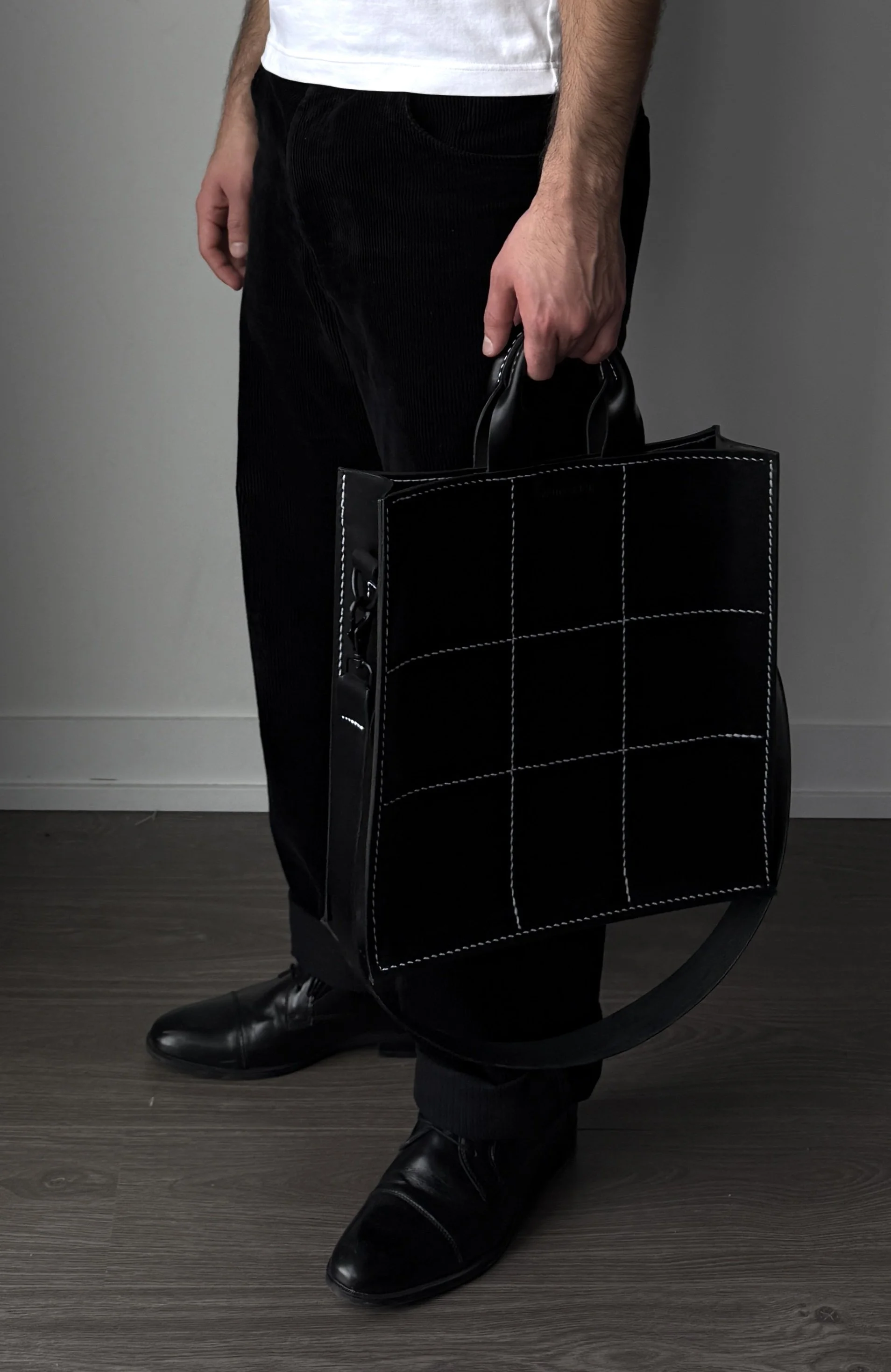 Person holding a large black tote bag with white stitching, dressed in black pants and shiny black shoes.