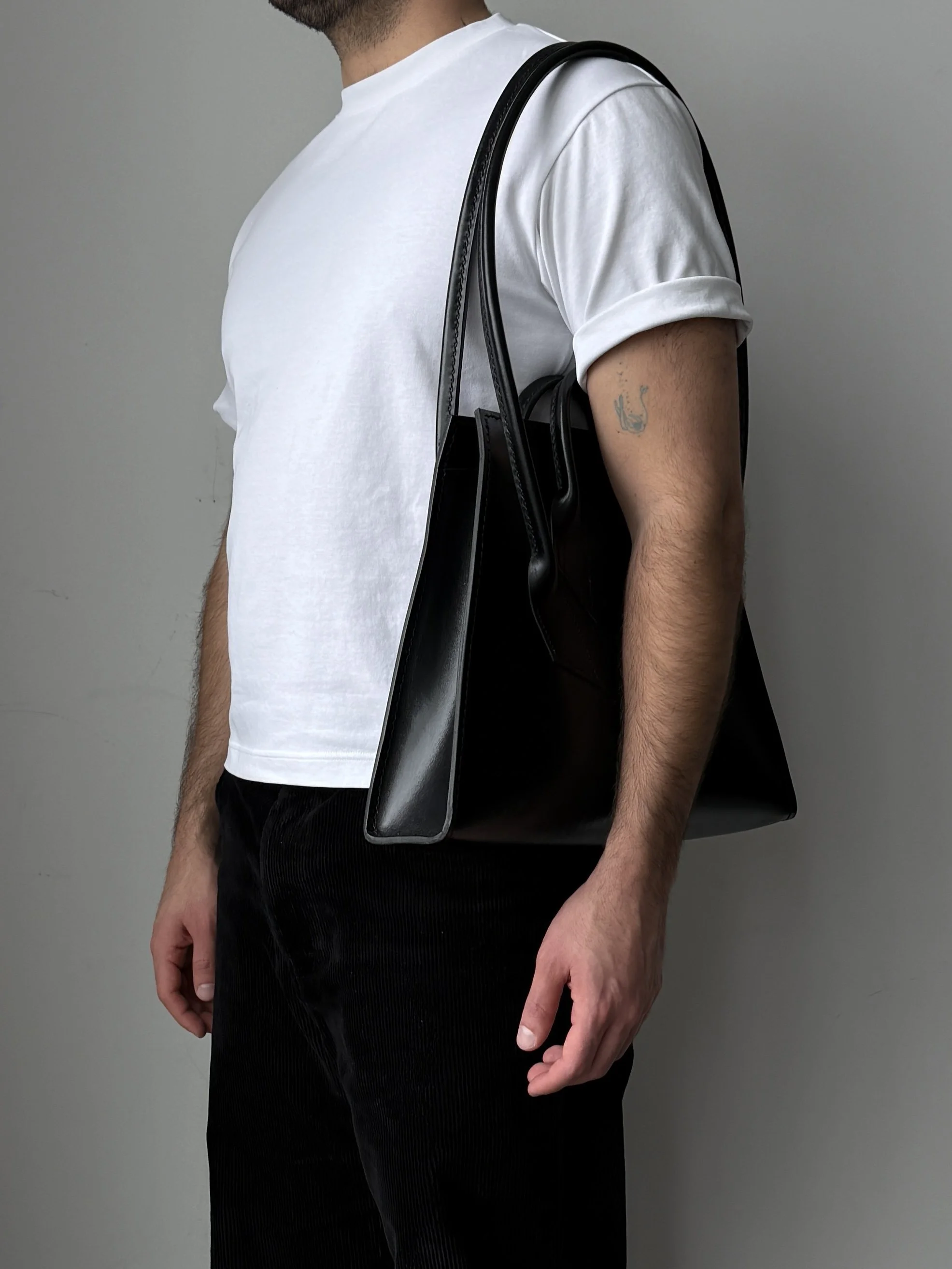 A man wearing a white t-shirt and black pants with a black shoulder bag over his shoulder, standing against a plain gray background.