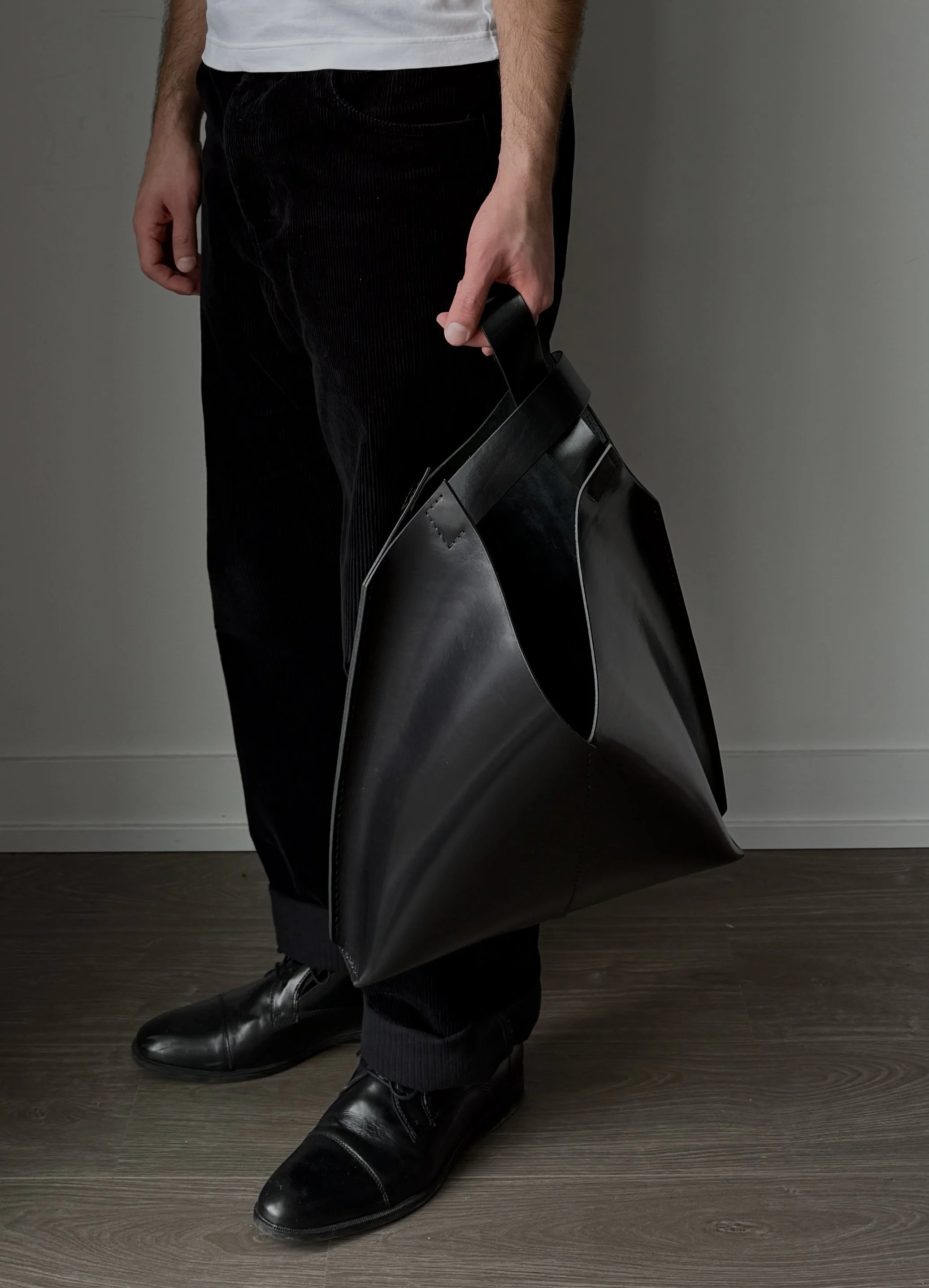 Person standing on a wooden floor holding a large black bag, dressed in black pants and black shoes.