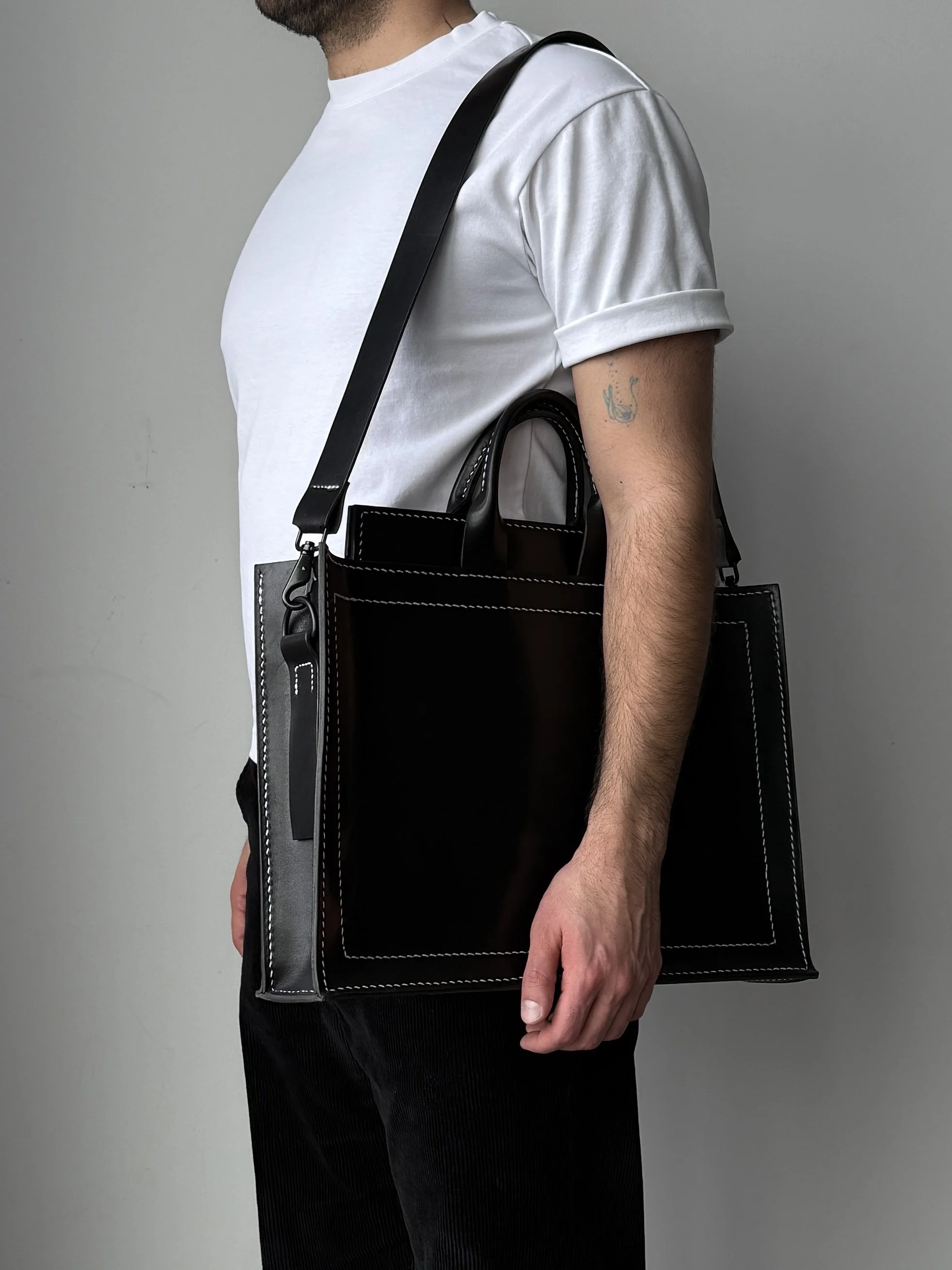 A person is wearing a white t-shirt and black pants, carrying a black bag with white stitching on the shoulder.