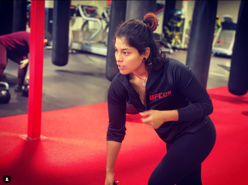 A woman with dark hair tied in a bun, wearing a black UFC Gym jacket, is exercising on a red mat at a gym with punching bags and workout equipment in the background.