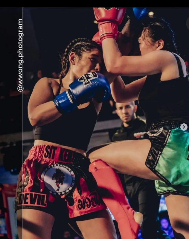 Two women fighters engaged in a mixed martial arts fight inside a ring, one wearing blue gloves and red shorts with black and pink design, the other wearing red gloves and green shorts, with a referee observing in the background.