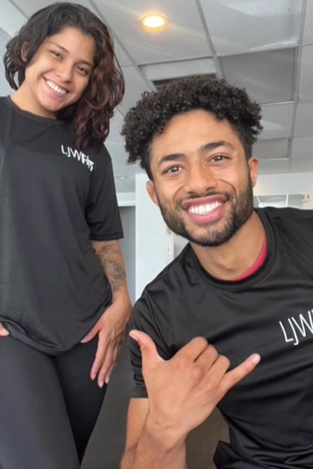 A man and woman smiling, wearing black shirts with white text, in an indoor setting with a ceiling and lights visible.
