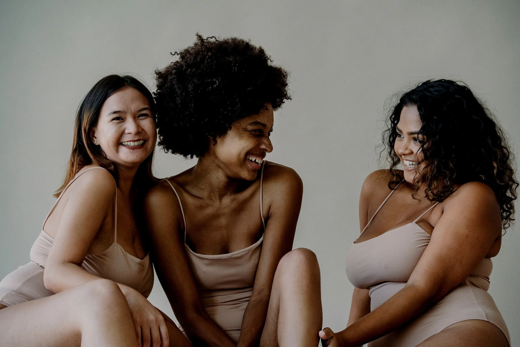 Three women in beige lingerie smiling and laughing together against a plain background.