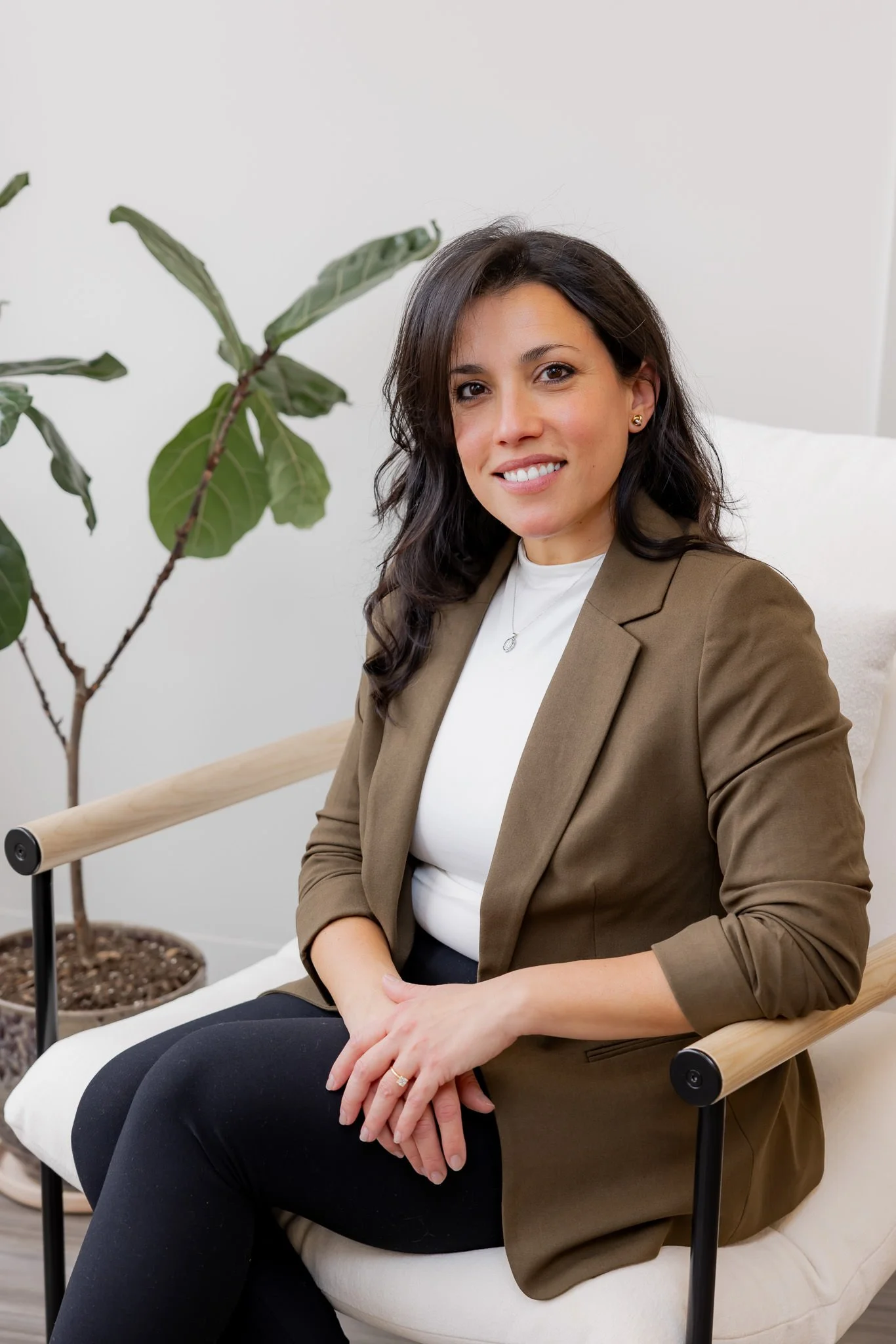 Nicole Dayton, Business Operations Manager at Morph Med Spa, seated in a modern chair wearing an olive blazer, smiling warmly in a bright clinical office setting.