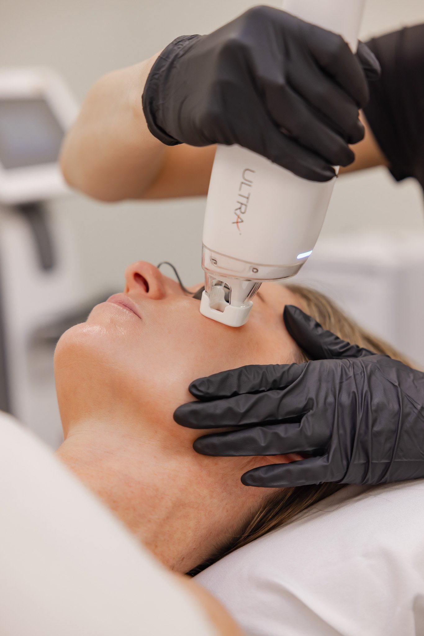 A woman receiving a facial treatment with a handheld skincare device while lying down in a spa or clinic. The technician is wearing black gloves.