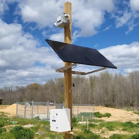 A wooden utility pole with a solar panel, a box-shaped device, and a satellite dish in an open field with a partly cloudy sky.