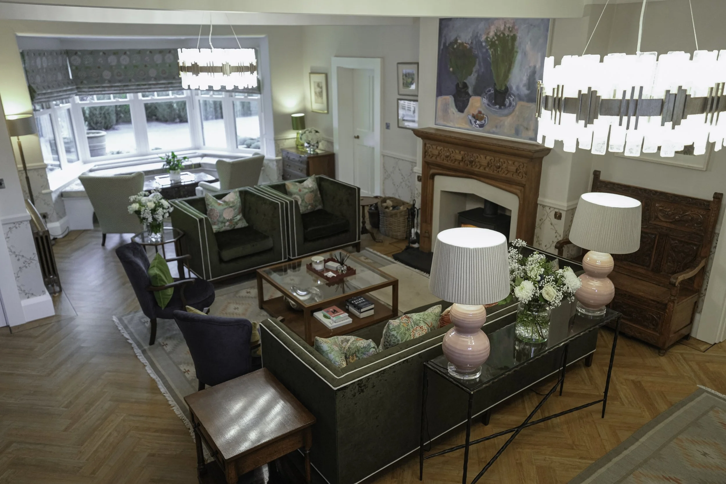 Living room with white paneled walls, a dark green sofa with assorted pillows, a modern black and wooden coffee table, a patterned rug, and a black bookshelf with decorative items and books, illuminated by natural light from a large window.