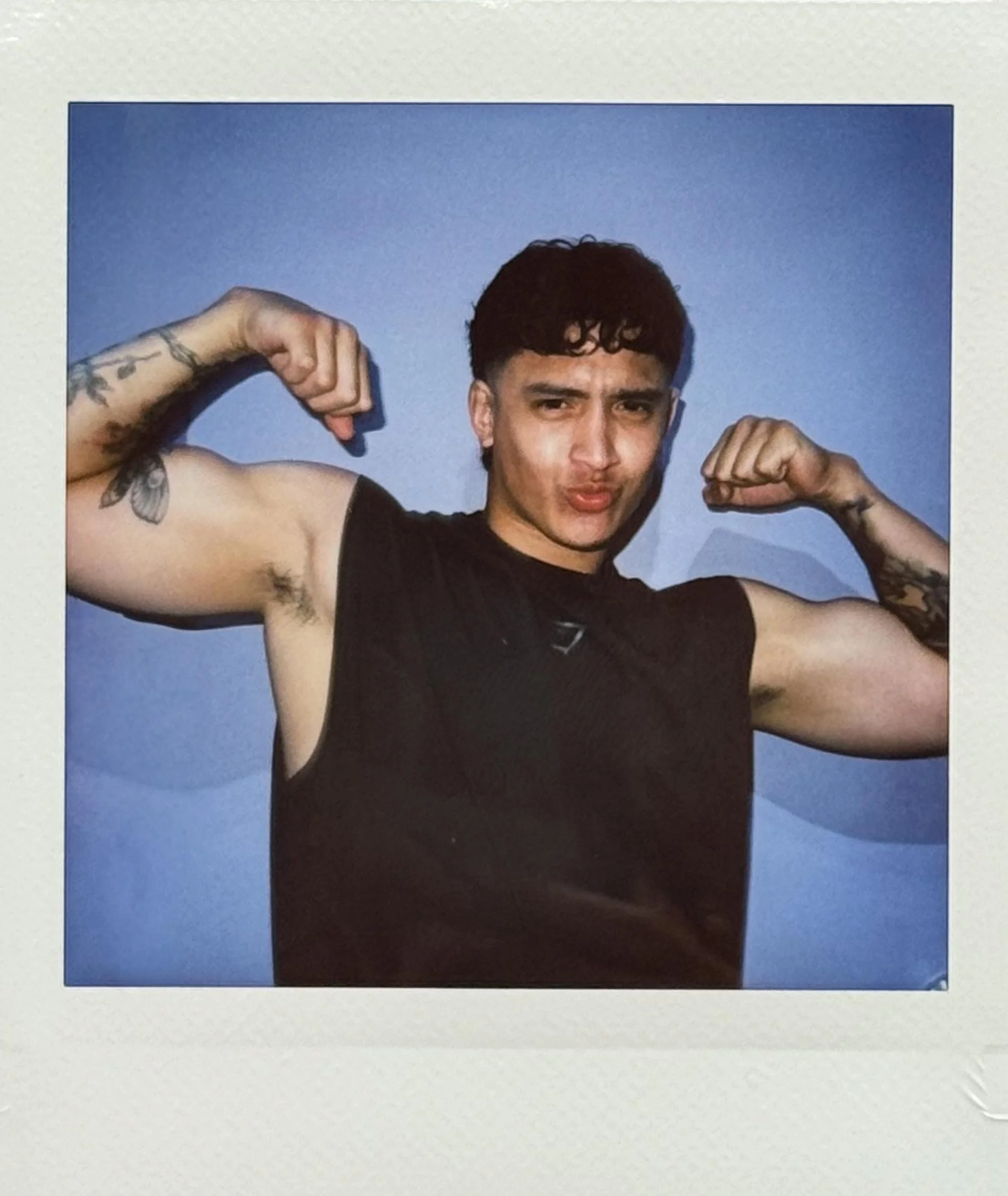 Young man with tattoos flexing muscles, making a playful face, wearing a black sleeveless shirt, against a blue wall.