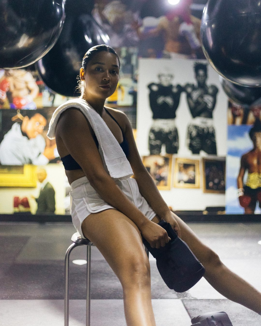 A woman sitting on a chair, wearing athletic wear and a towel around her neck, in a gym with boxing posters and photos in the background.