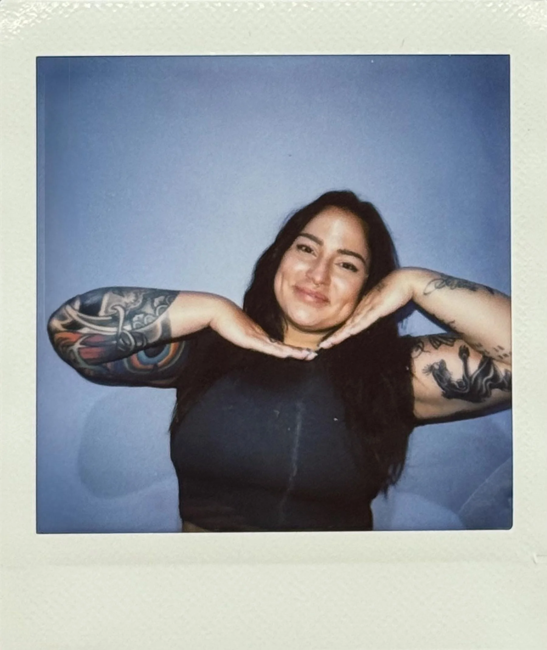 A woman with tattoos on her arms, smiling, with her hands under her chin, against a blue background.