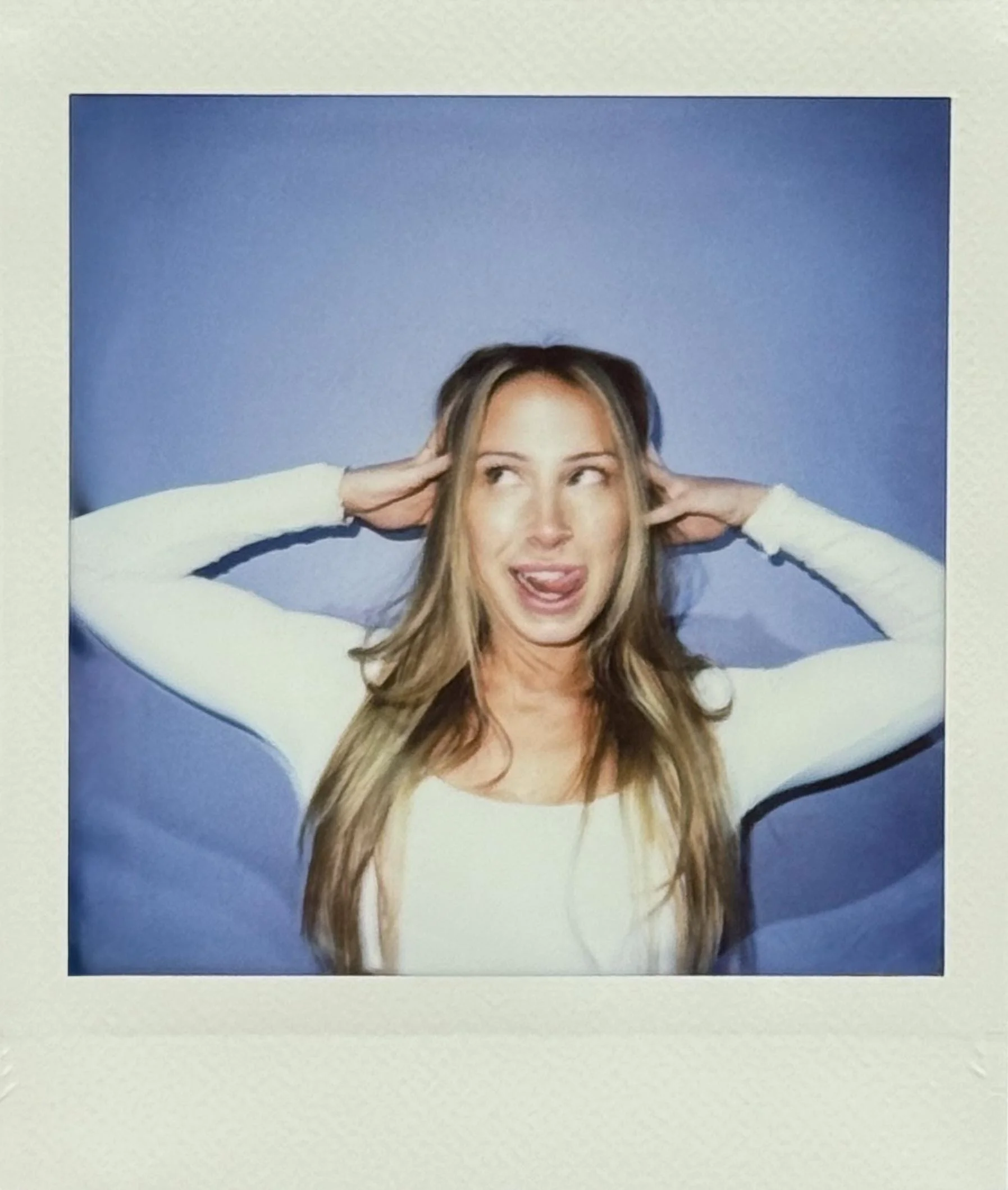 A woman with long blonde hair making a silly face, sticking her tongue out and crossing her eyes, while holding her hands behind her head. She is wearing a long-sleeved white top and standing against a blue background.