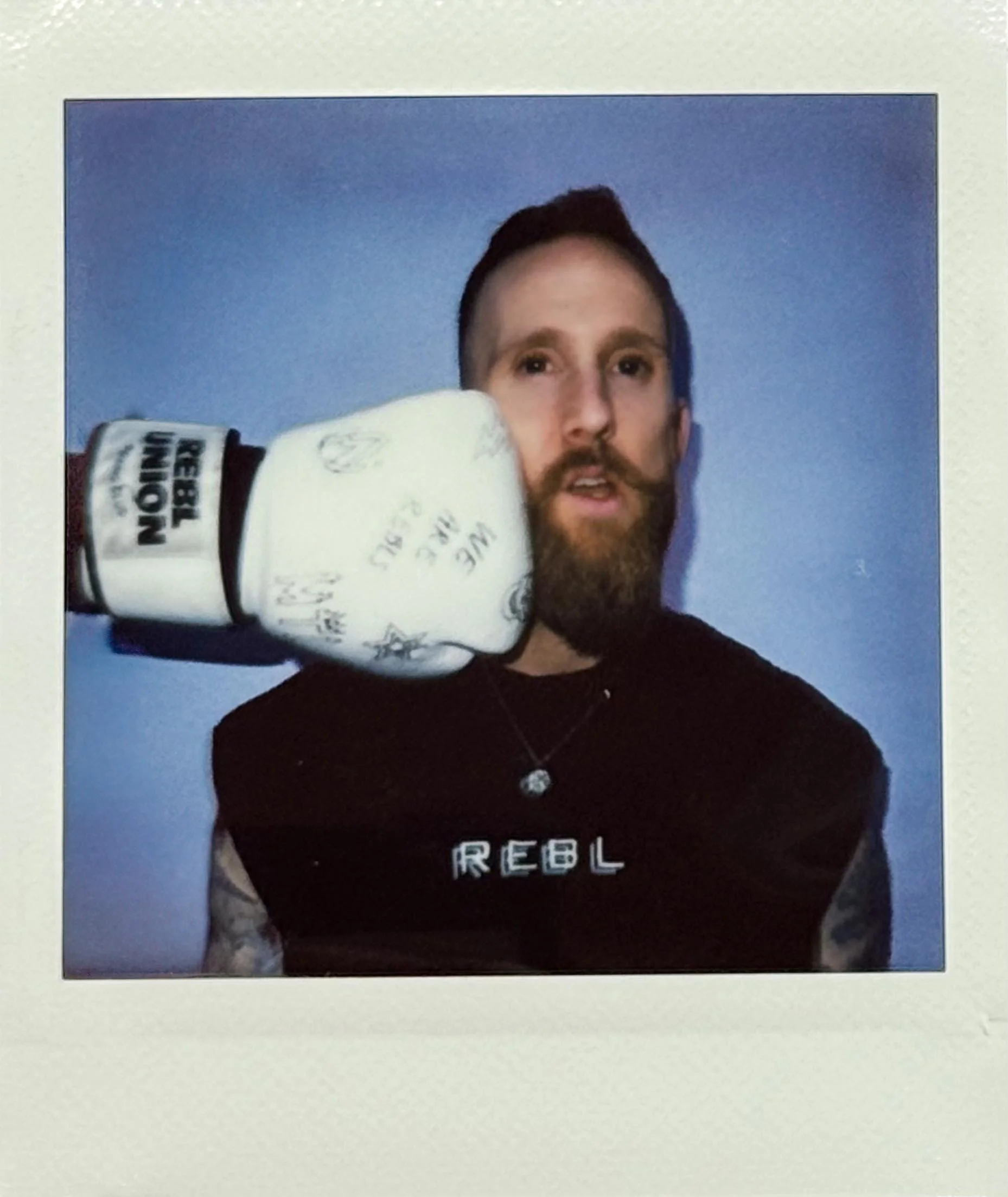 A man with a beard and mustache wearing a black shirt with the word 'REBL' on it and a necklace, being punched in the face by a hand wearing a white boxing glove with signatures on it, against a blue background.