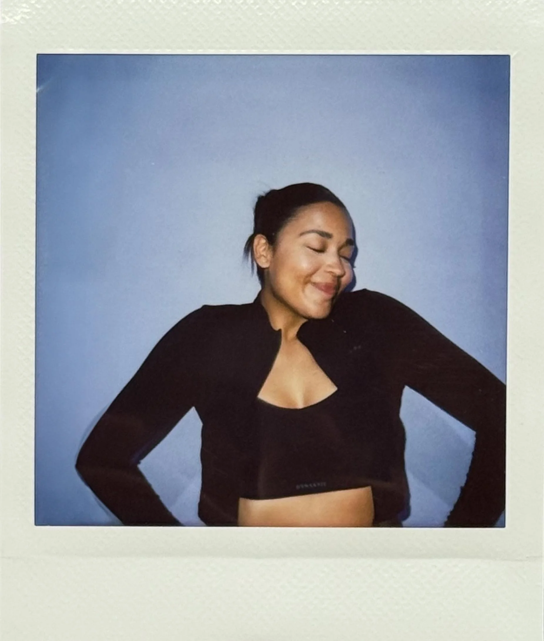 Young woman smiling with her eyes closed, wearing a black crop top and black jacket, against a blue background.