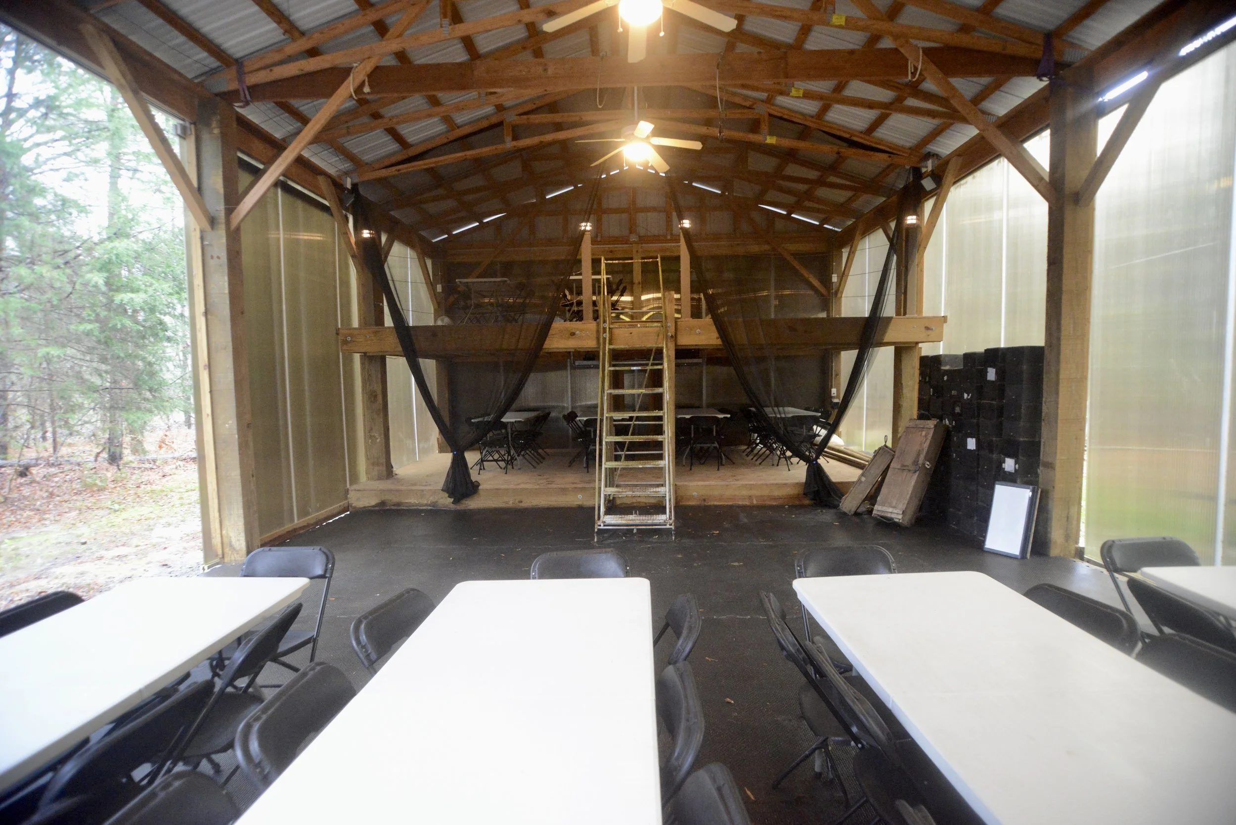 Inside a pavilion with wooden beams, metal roof, and large tables with folding chairs. A loft area with stairs and netting is visible. Outdoors can be seen through semi-transparent walls.