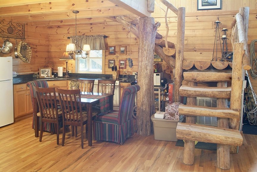 Rustic cabin interior with wooden staircase, dining table, chairs, and kitchen area. Walls are made of logs, and the space has a cozy, homey feel.