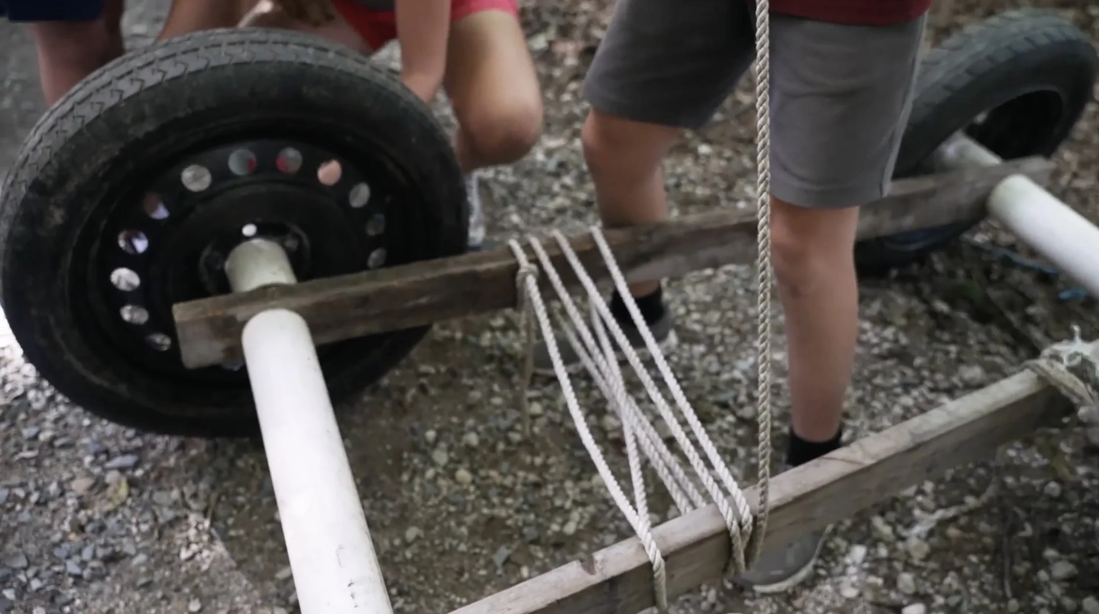 Two people assembling a wooden and metal cart with rope and wheels on a gravel surface.