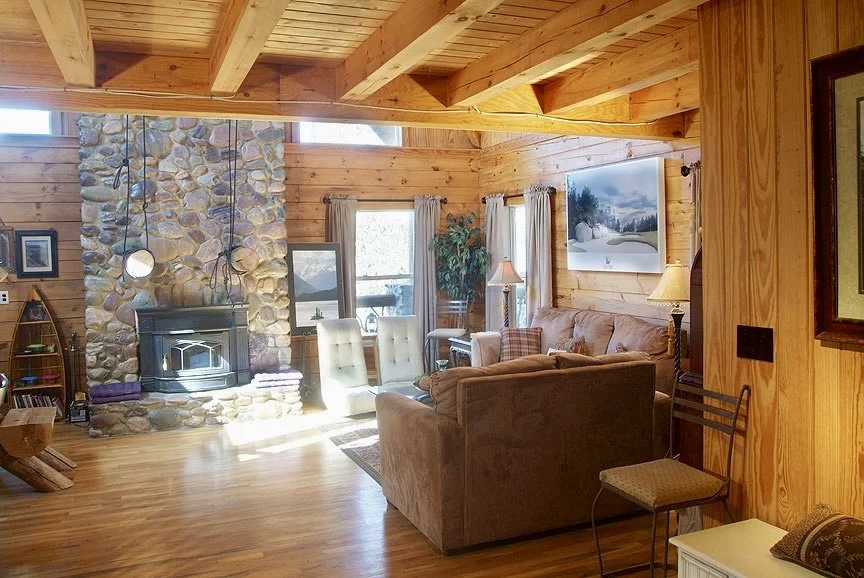 Cozy log cabin living room with wooden walls and ceiling, stone fireplace, plush sofas, and natural light streaming in from large windows.