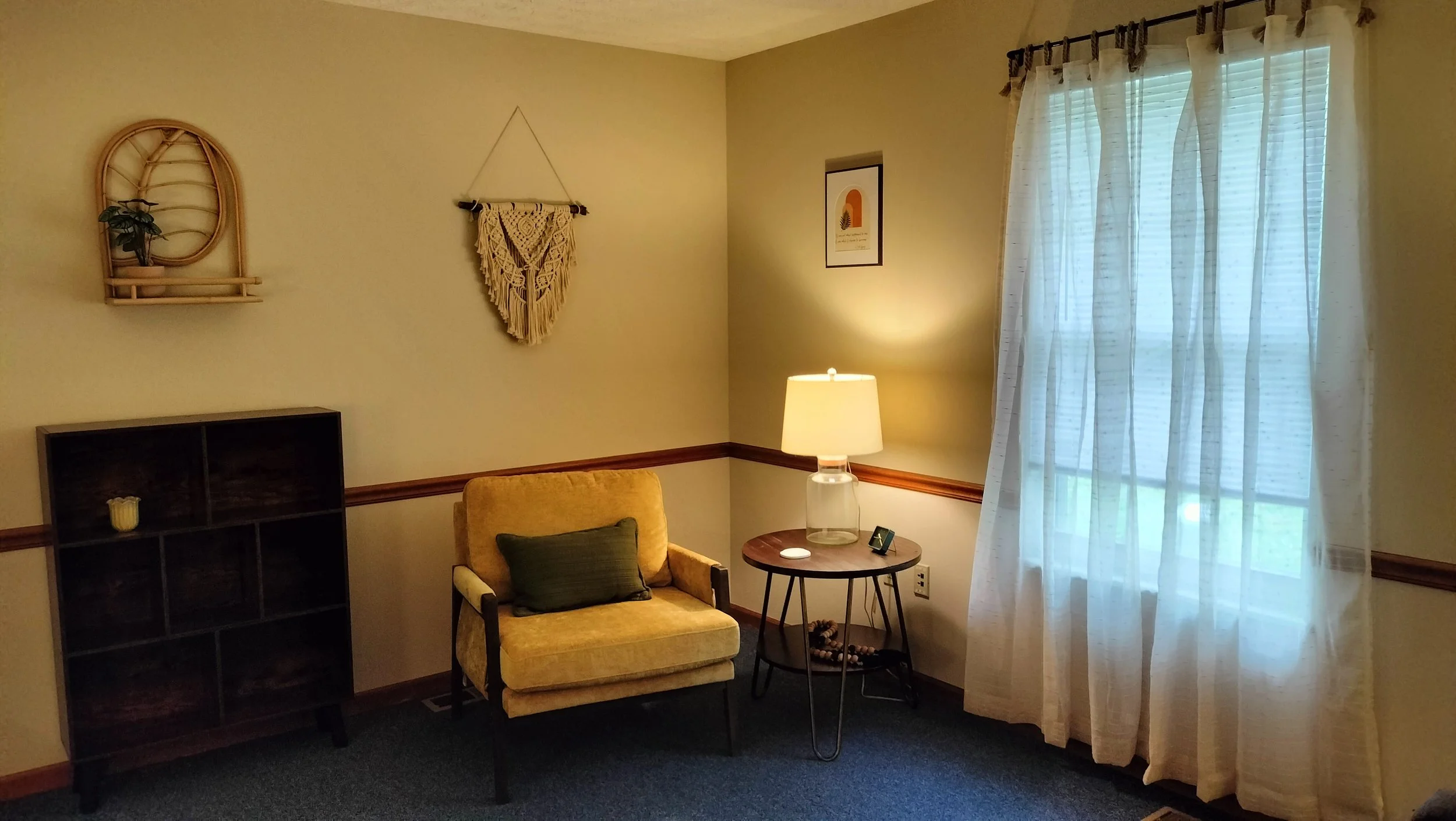 Therapist's office showing a bookshelf, chair, side table with lamp, window, and art on the wall