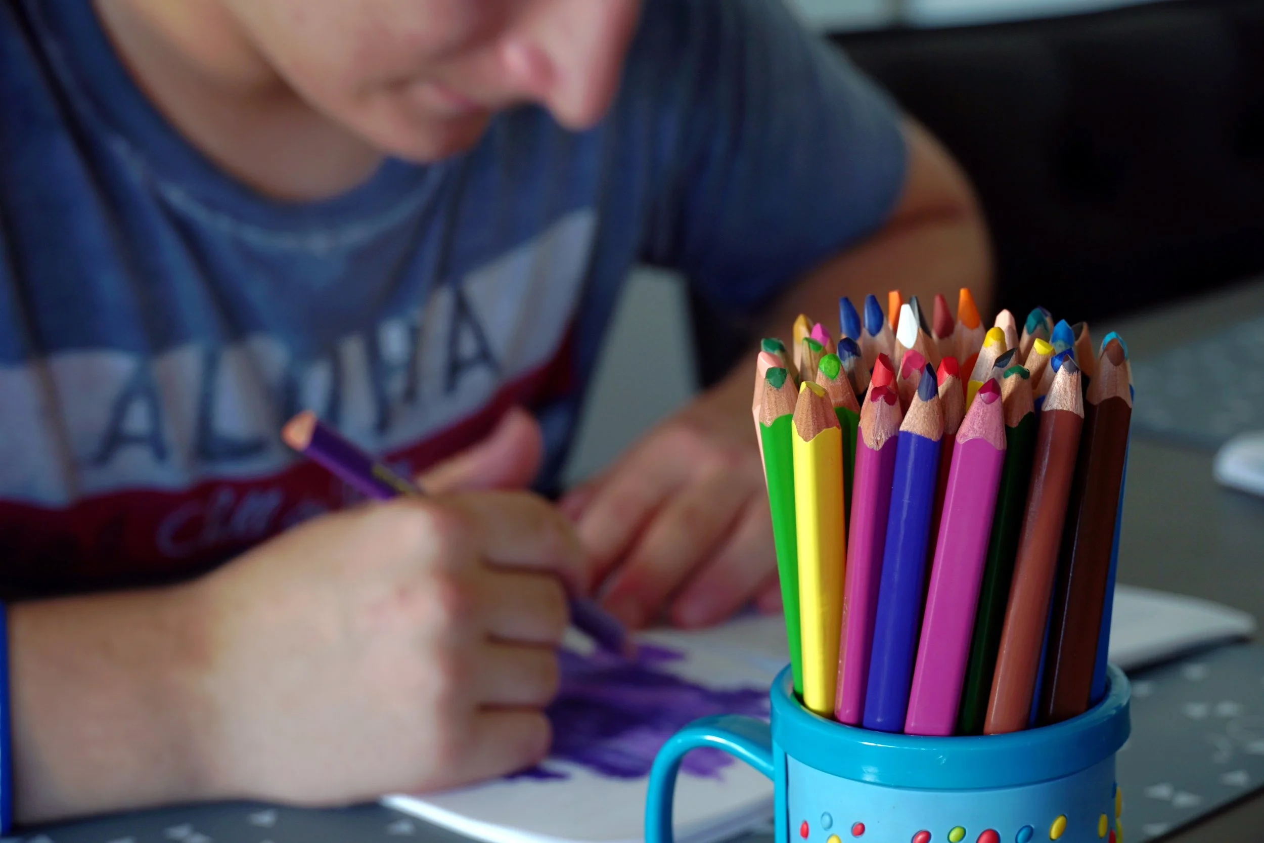 teen boy coloring with colored pencils.  There is a pad of paper and a cup of colored pencils nearby