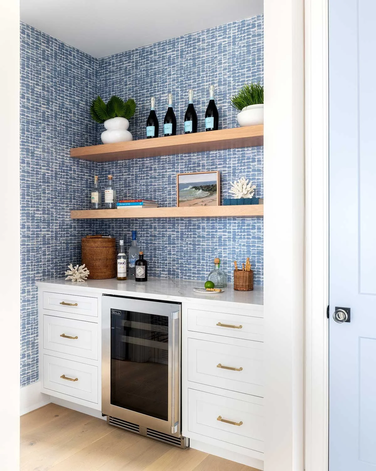 A bar area with white cabinets, a small wine fridge, and two wooden shelves on a blue mosaic wall, decorated with bottles, plants, books, and coastal decor.