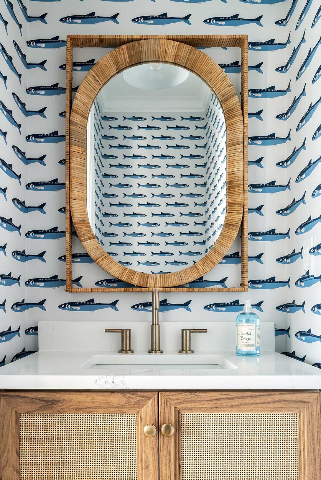A bathroom with a mirror framed by a wooden border, a fish-patterned wallpaper, a white sink with gold fixtures, a wooden cabinet with rattan panels, and a bottle of hand soap on the counter.