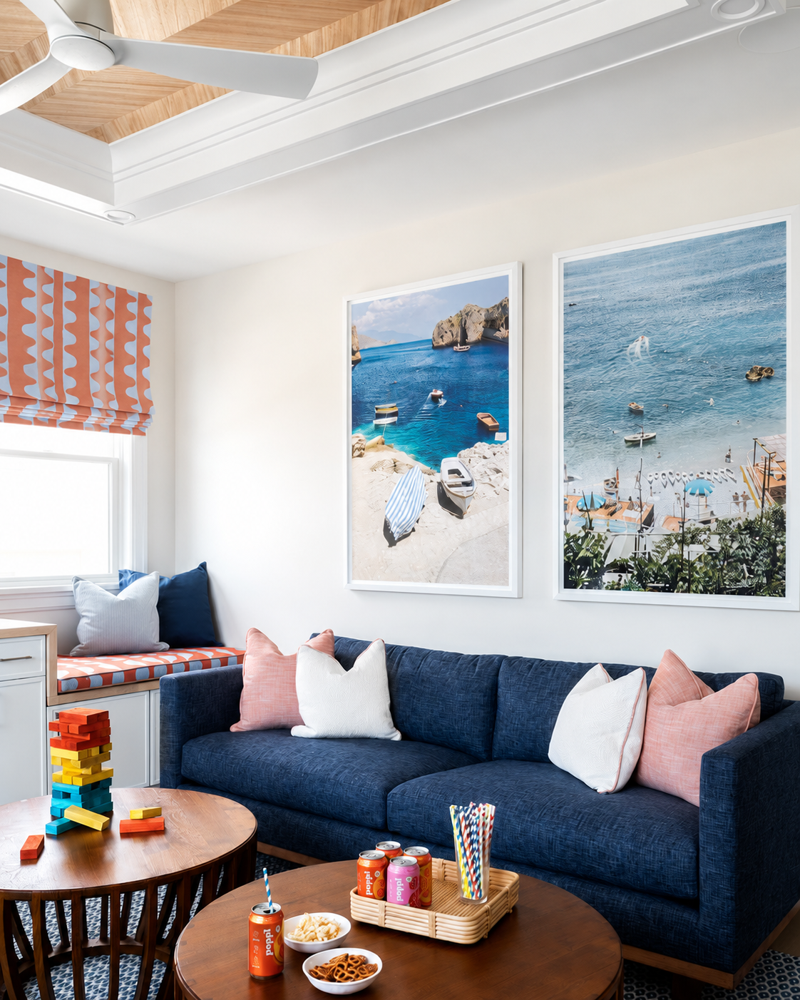 Living room with a navy sofa, pastel pillows, a wooden coffee table with snacks and drinks, a built-in window seat with pillows, and large framed beach scene photographs on the wall.