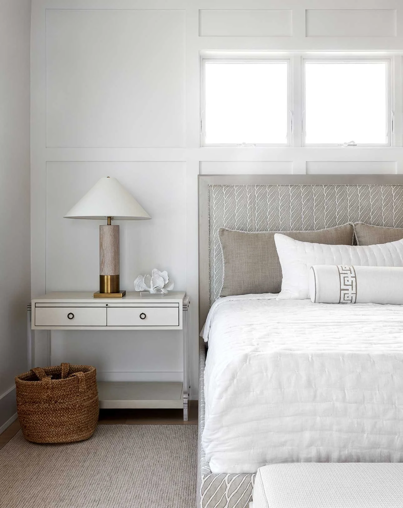 A bedroom with a white bed, gray headboard, nightstand with a lamp, coral decoration, and a woven basket, with a window in the background.