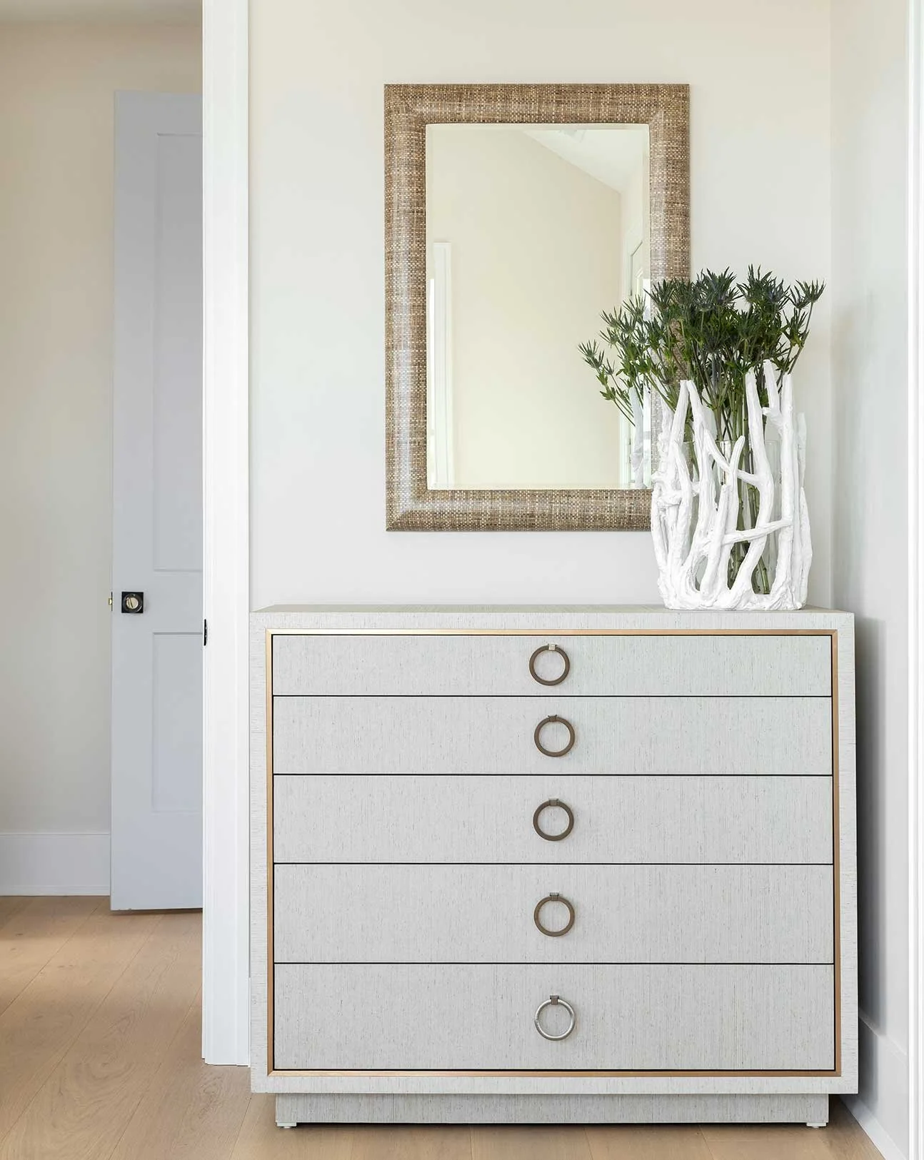 White dresser with circular handles, a large mirror with a woven frame above it, and a white floral arrangement with green foliage in a white decorative vase on a light-colored wooden floor.