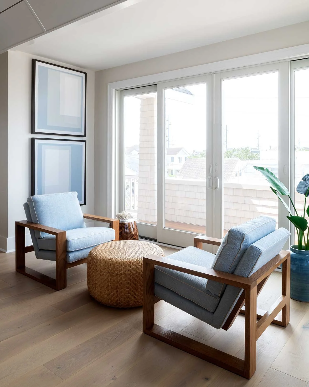 A bright living room with two blue upholstered armchairs with wooden frames, a round wicker ottoman, large sliding glass doors leading to a balcony, framed abstract art on the wall, and a potted plant in a blue ceramic pot.