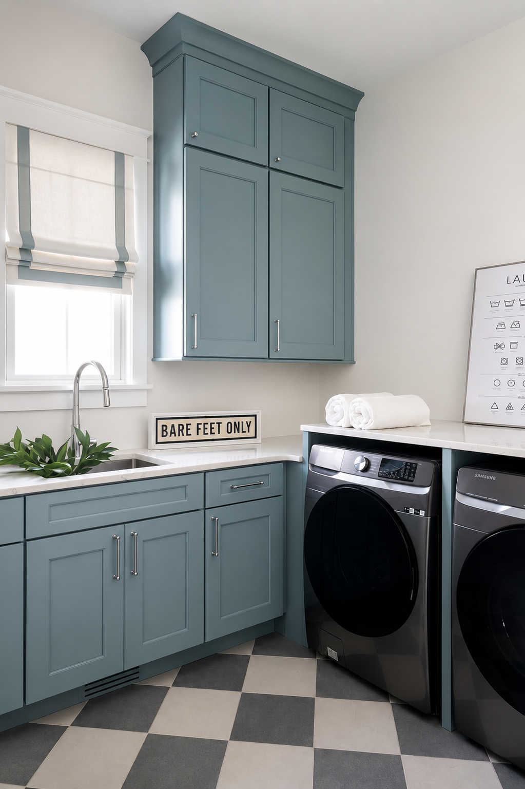 A laundry room with blue cabinets, a white countertop, a small sink with a plant, a sign that reads 'BARE FEET ONLY', a window, folded towels, a laundry chart, and two front-loading washing machines on the right.