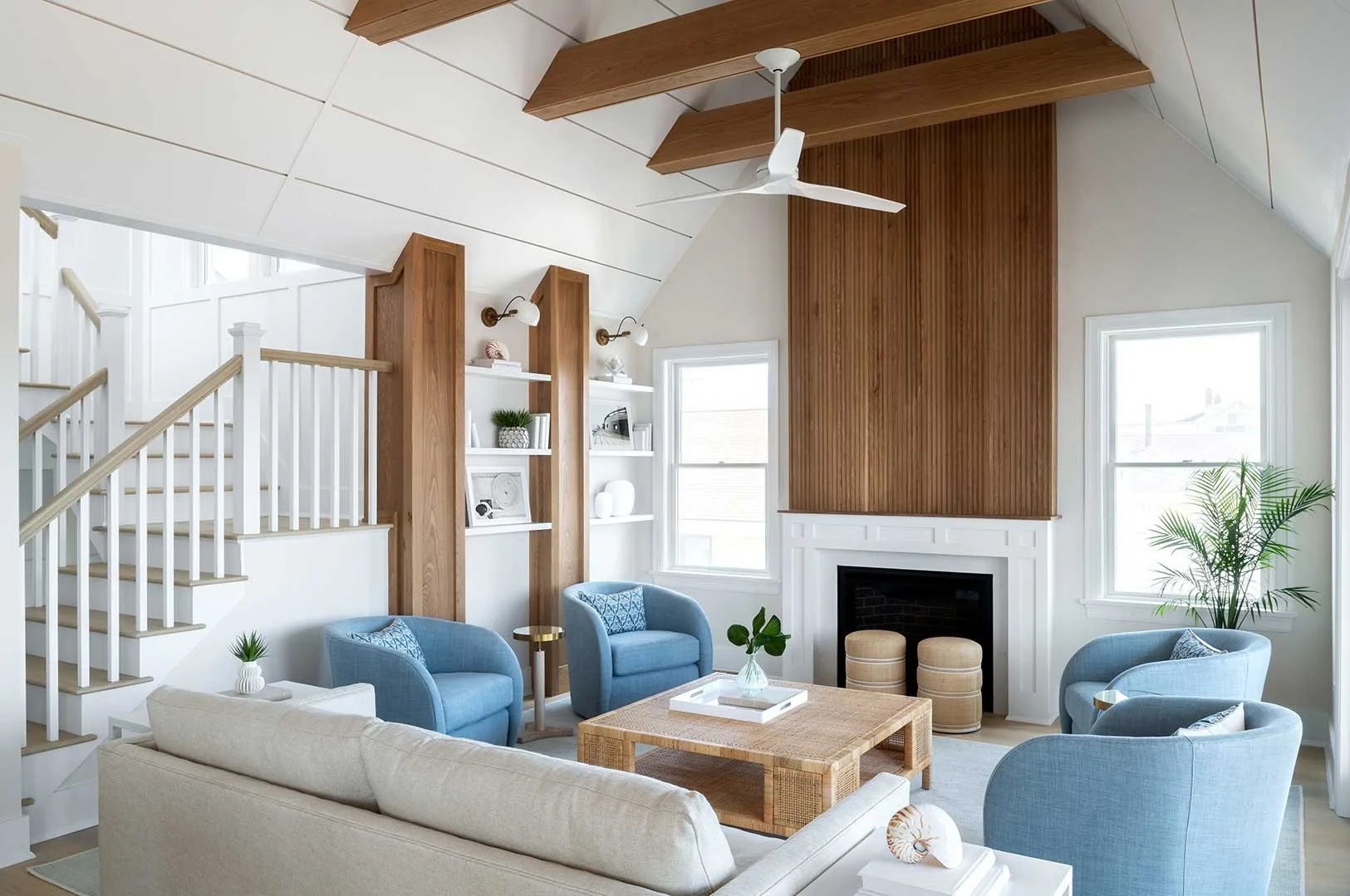 Living room with white walls, vaulted ceiling with wooden beams, blue armchairs, a beige sofa, a wooden coffee table, white built-in shelves with decor, a fireplace, large windows, and plants.