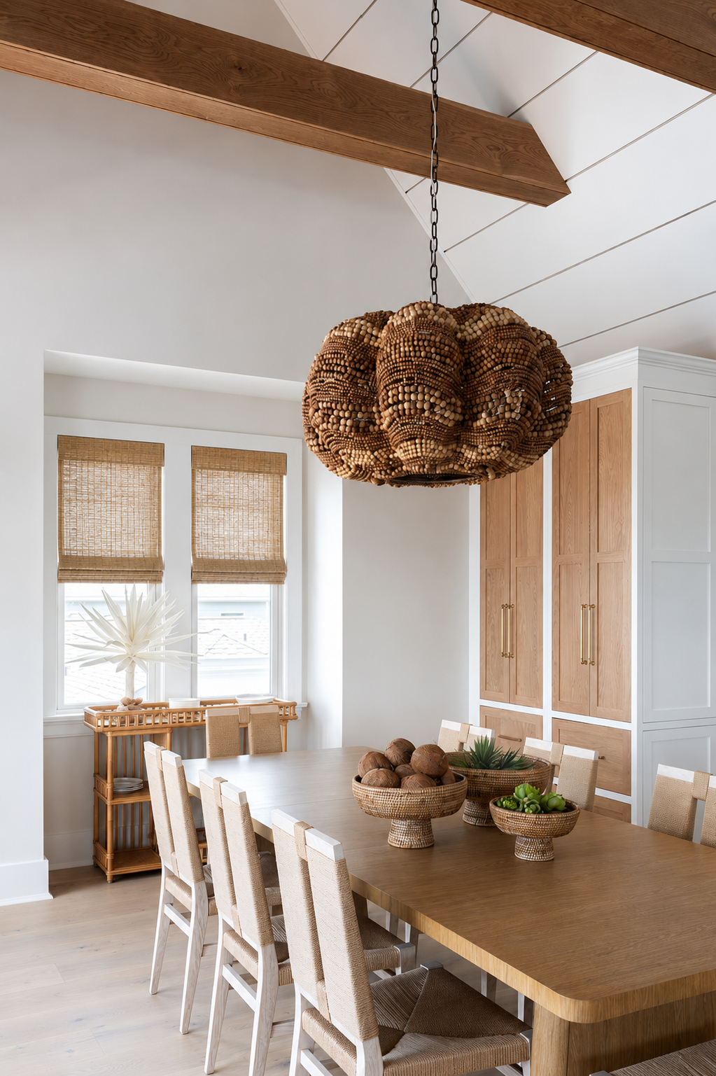 A dining room with a wooden table, beige chairs, a large pendant light made of wood beads, a basket with decorative brown balls, and a white window with natural light.