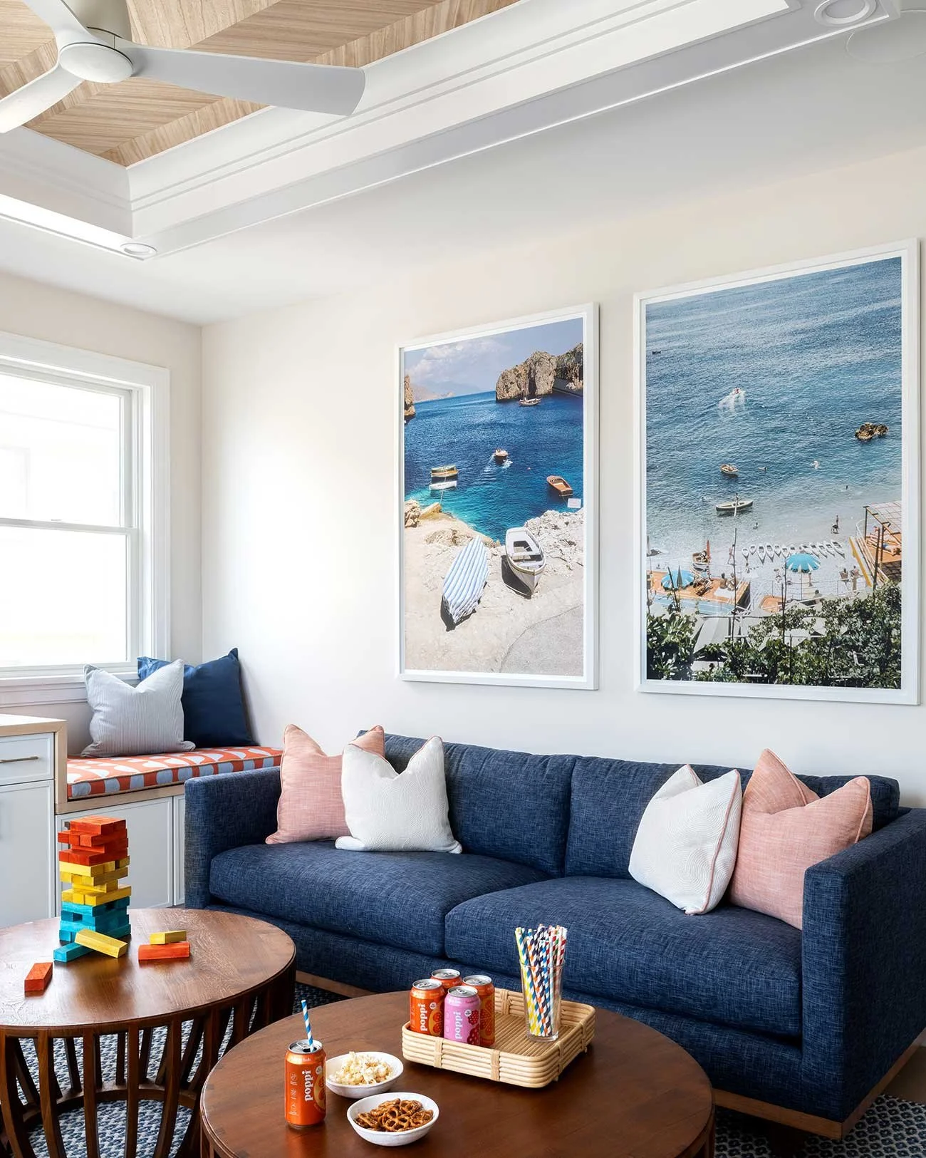 Living room with a navy sofa, pastel pillows, a wooden coffee table with snacks and drinks, a built-in window seat with pillows, and large framed beach scene photographs on the wall.