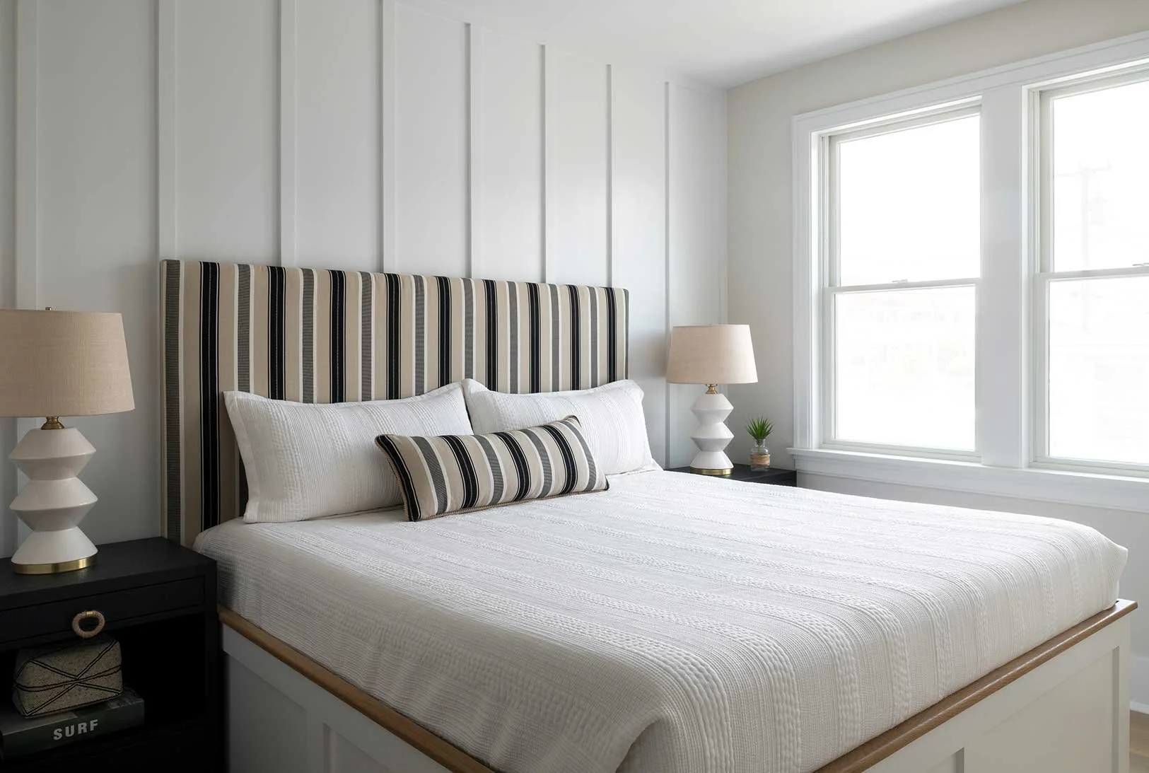 A neatly made bedroom with a large bed featuring a striped headboard, white bedding, and matching striped pillows. There are two nightstands with beige lamps, a small potted plant, and a window with white framing allowing natural light.