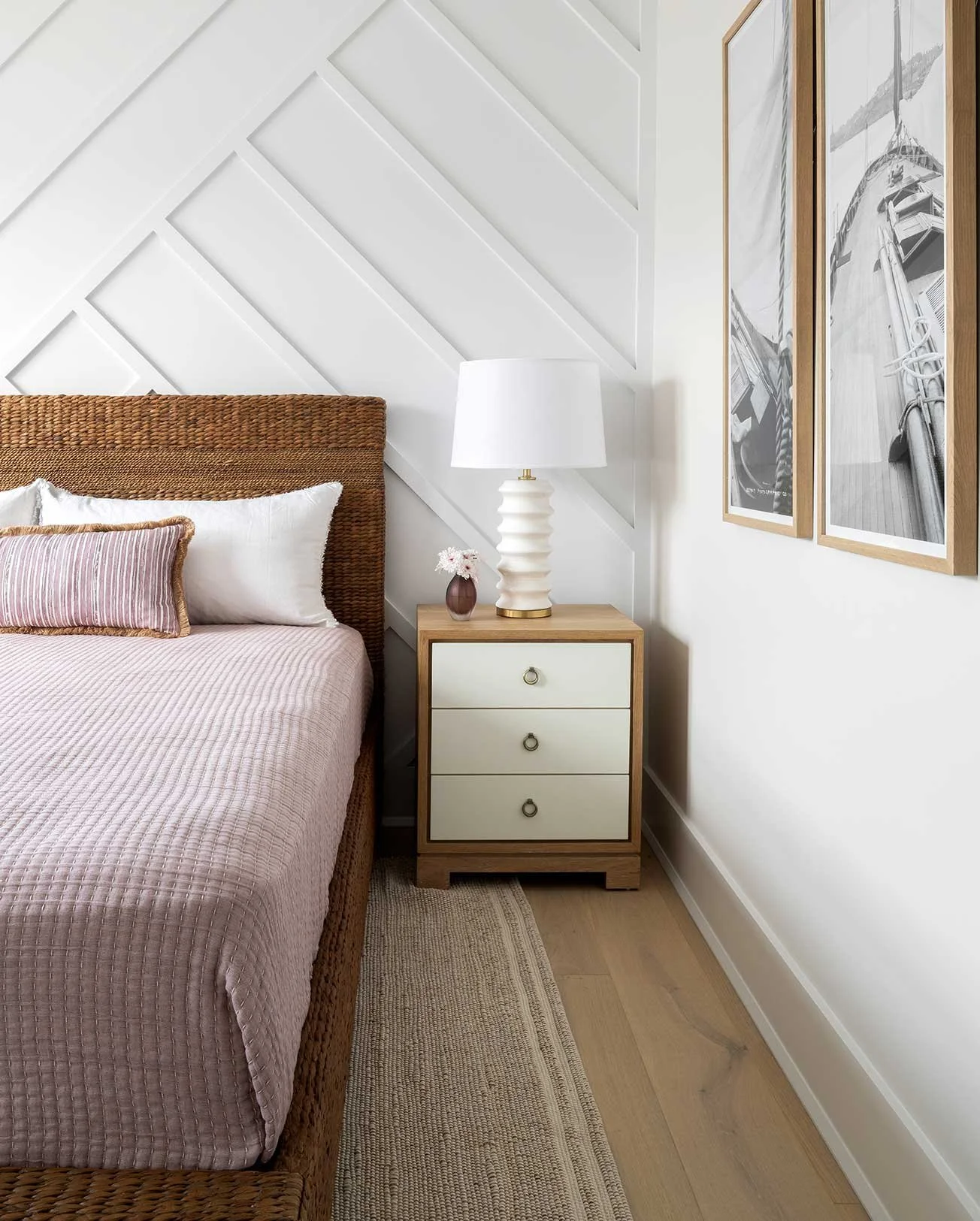A bedroom with a wooden bed frame, light pink quilt, white pillows, and a pink striped pillow. A wooden nightstand with three drawers, a white ceramic lamp, and a small vase with flowers. Two framed black and white photos hang on the white wall next 