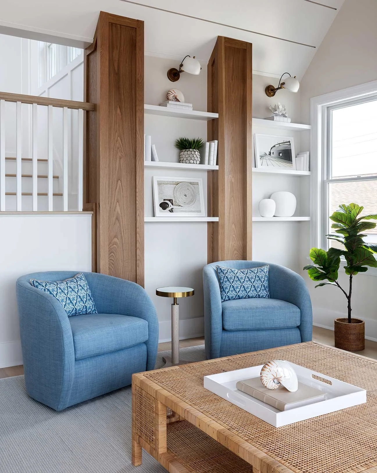 Living room with two blue armchairs, a wicker coffee table with a decorative tray and seashell, a small side table, a tall green plant, and built-in white shelves with decorative items and framed photos, next to a large window.