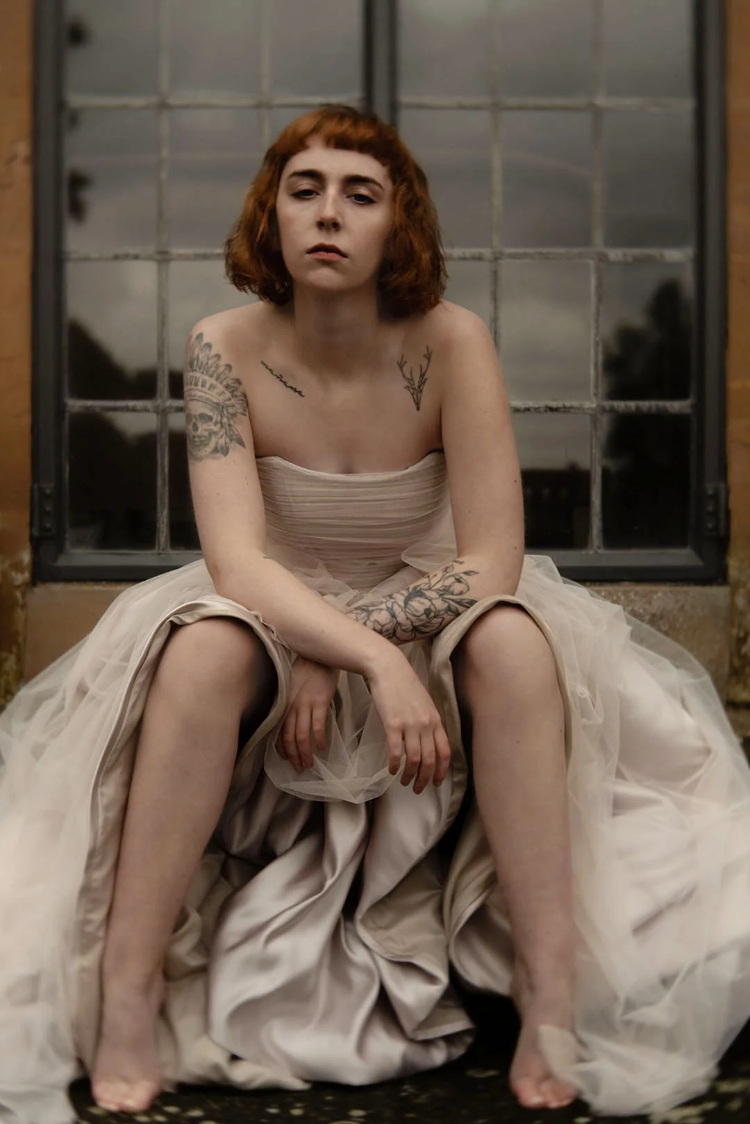 Red-haired-tattoed-young-woman-sitting-by-a-window-in-a-wedding-dfress