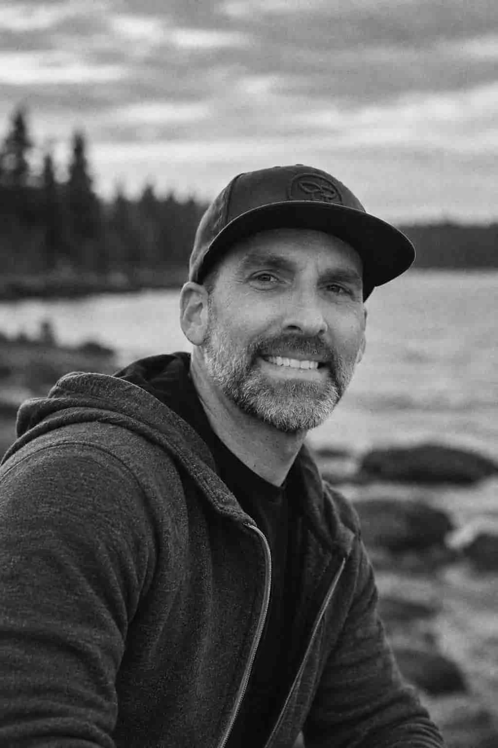 A man with a beard and mustache smiling at the camera, wearing a cap and a zip-up hoodie, outdoors by a body of water with trees and cloudy sky in the background.