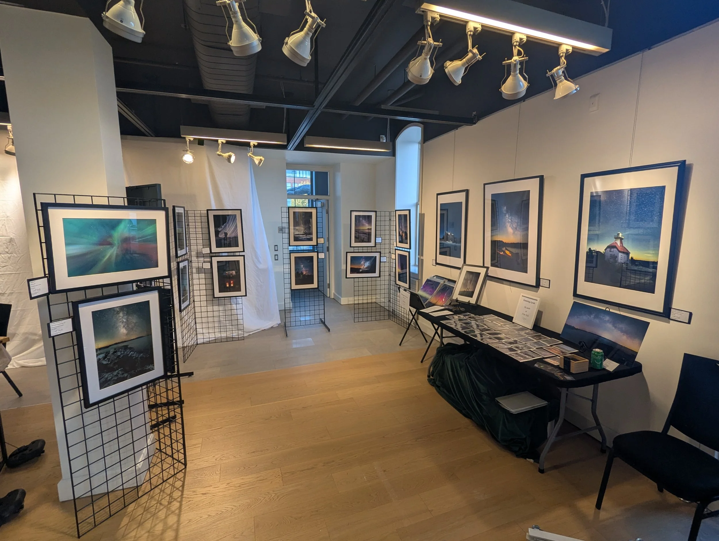 An art gallery showcasing framed landscape photographs arranged on black metal grid display panels and a table with art prints for sale. Track lighting illuminates the artwork, with a window and white curtain in the background.