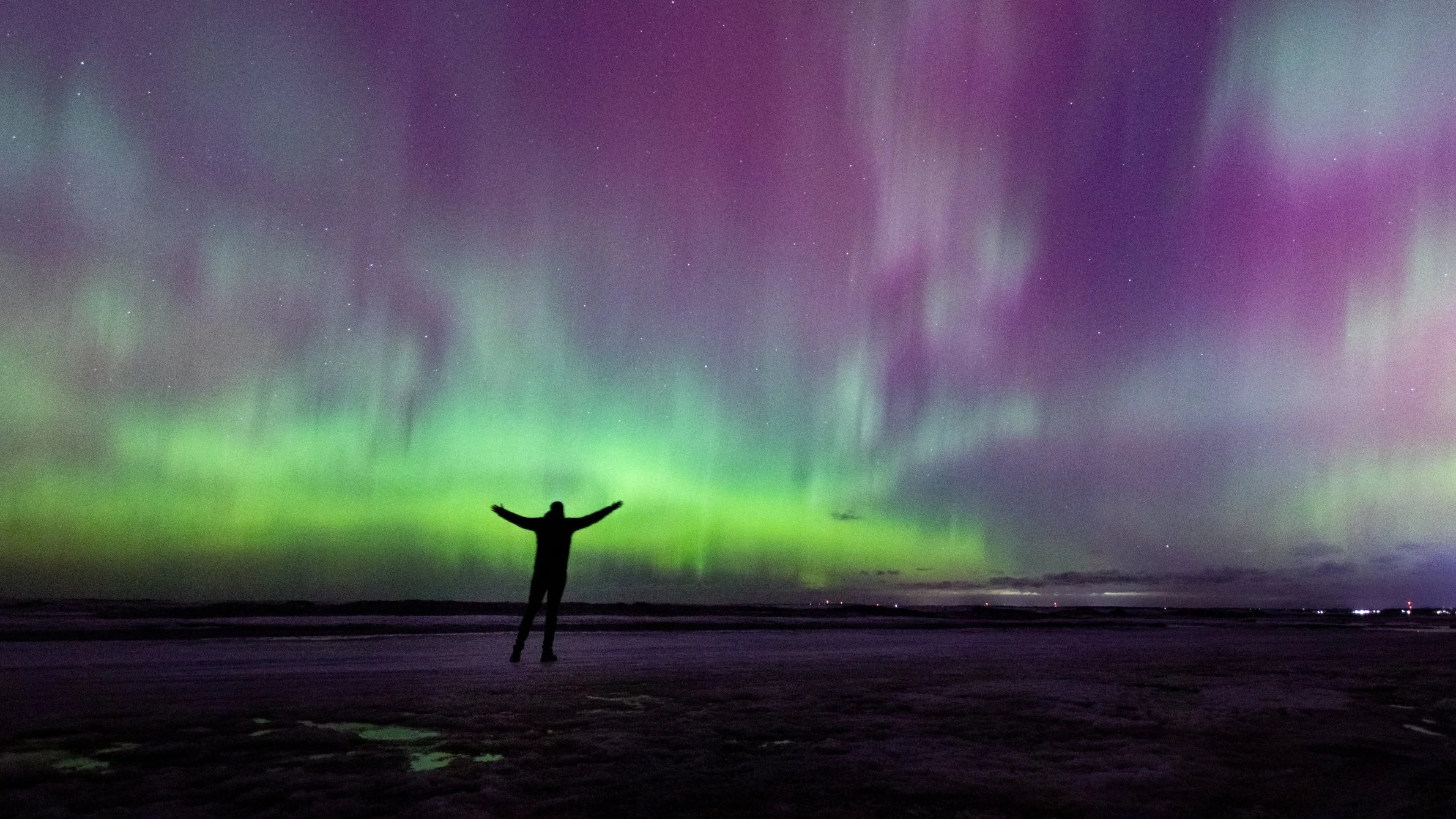 Person standing on a beach at night with arms raised under colorful northern lights in green, purple, and pink hues.