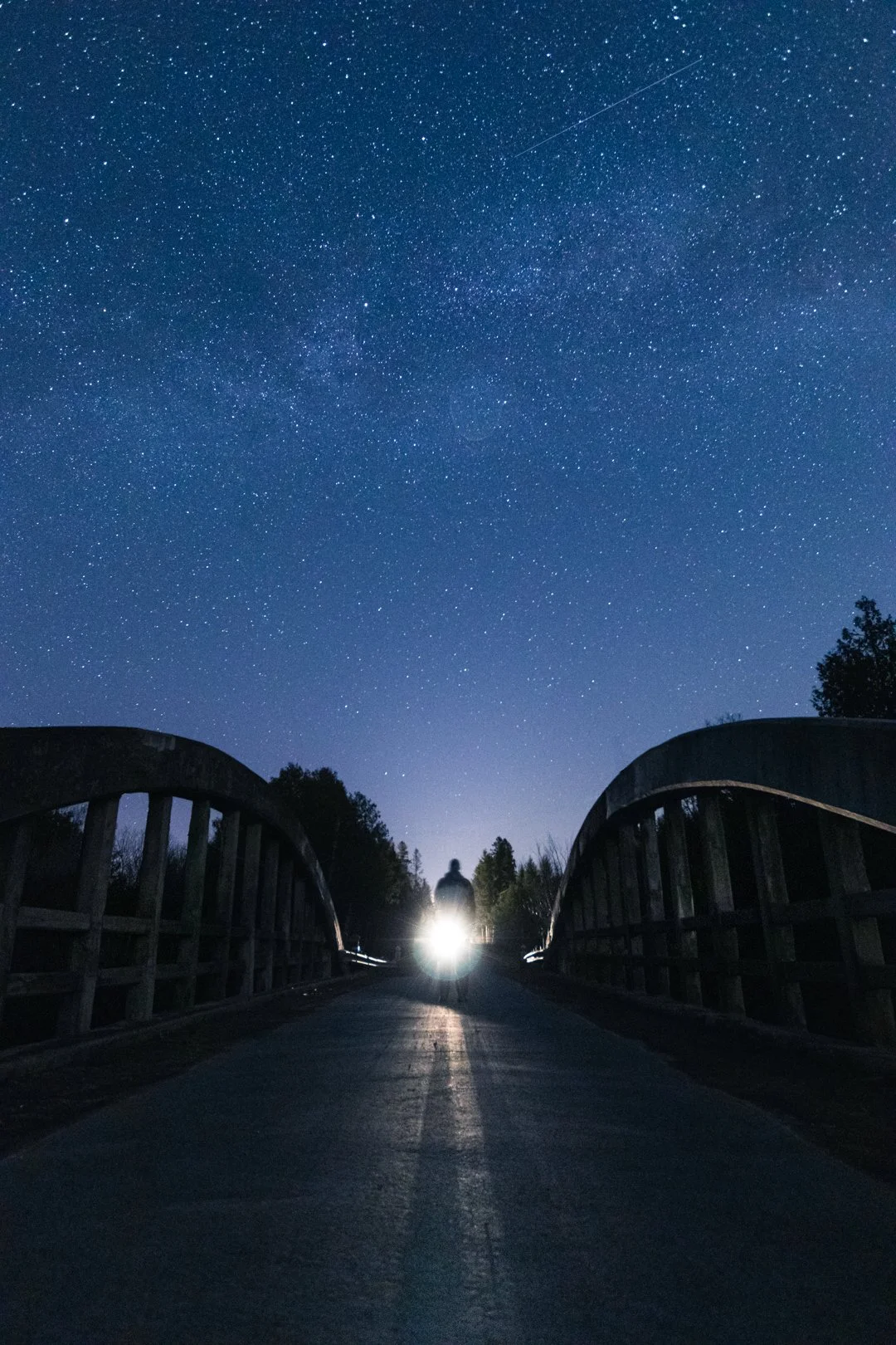 Person standing on a bridge at night illuminated by a bright headlight, with a starry sky and a shooting star overhead.