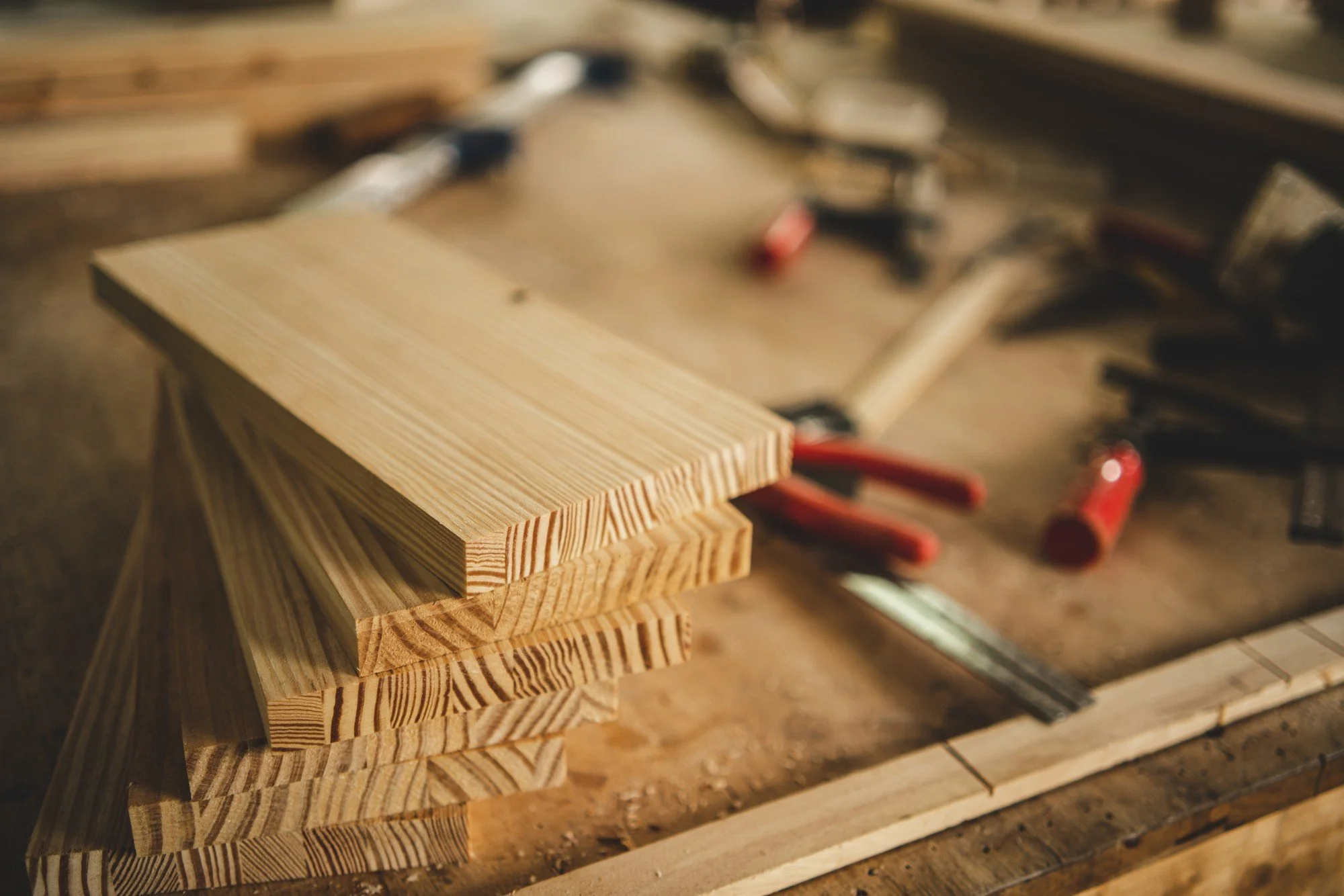 Stack of wooden planks on a workbench in a carpentry shop, with tools blurred in the background.