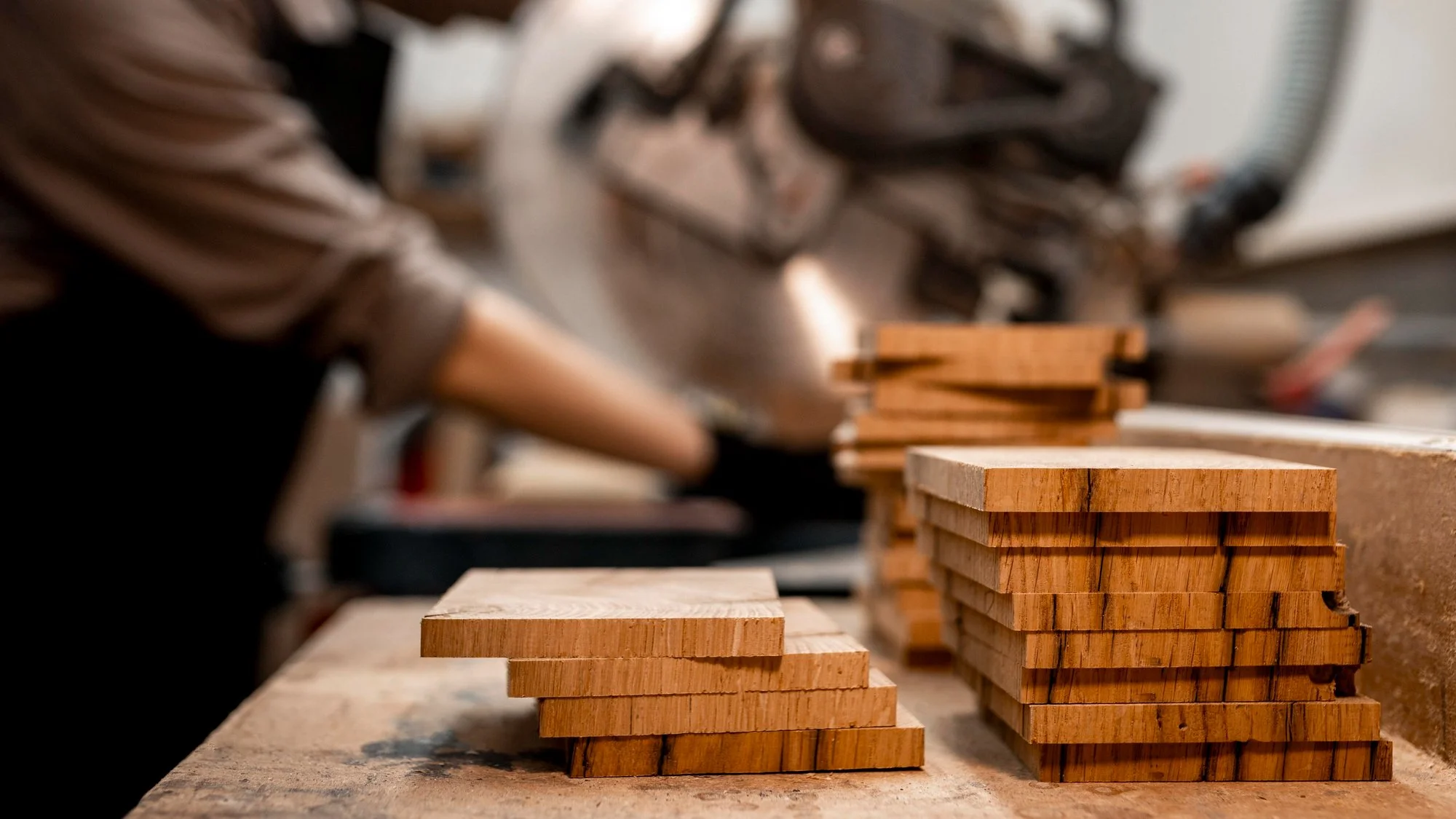 Stacks of cut wooden pieces on a workbench in a woodworking shop, with a blurred background of a person working.