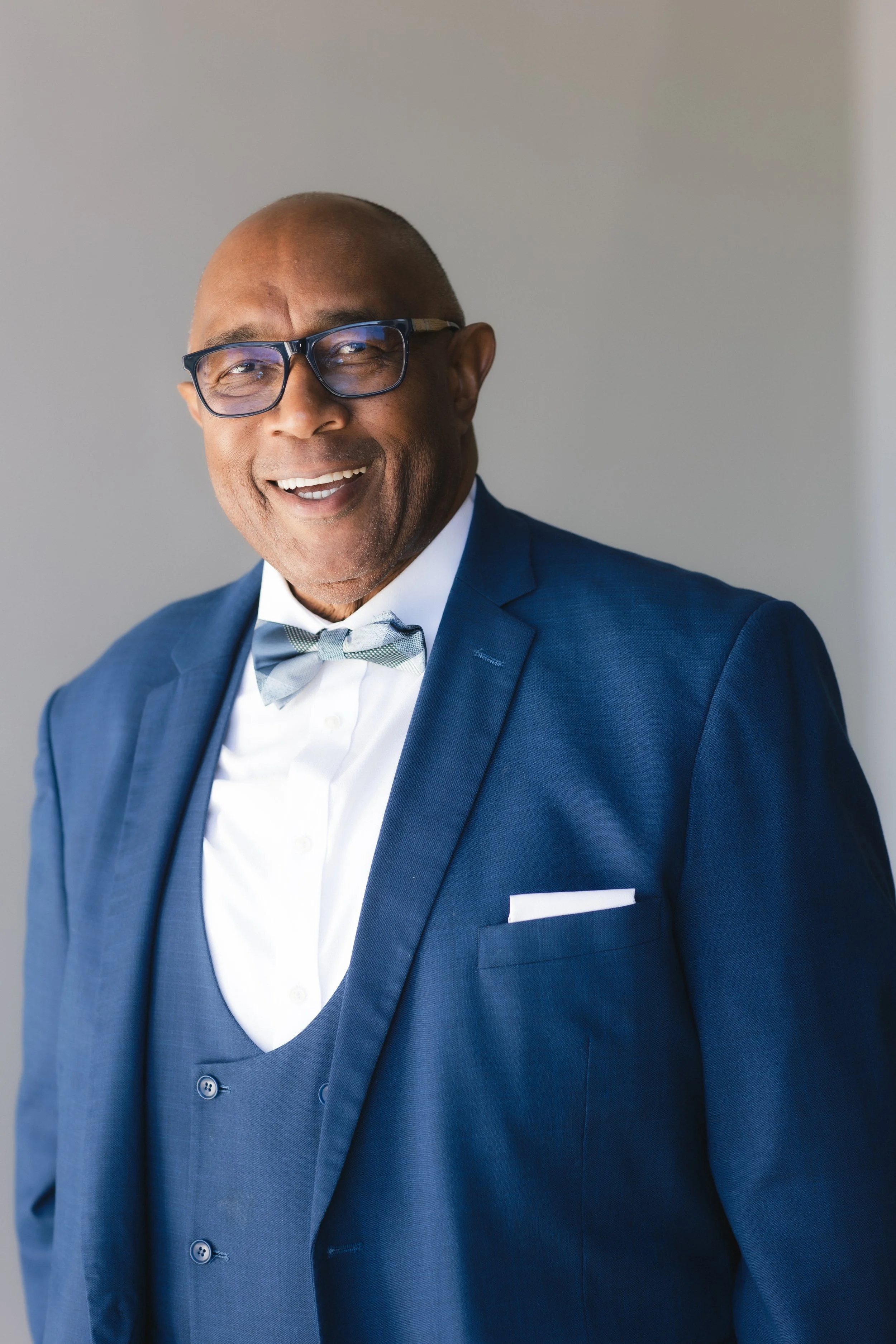 A smiling man wearing a dark blue suit, white shirt, gray bow tie, and glasses, standing against a neutral background.