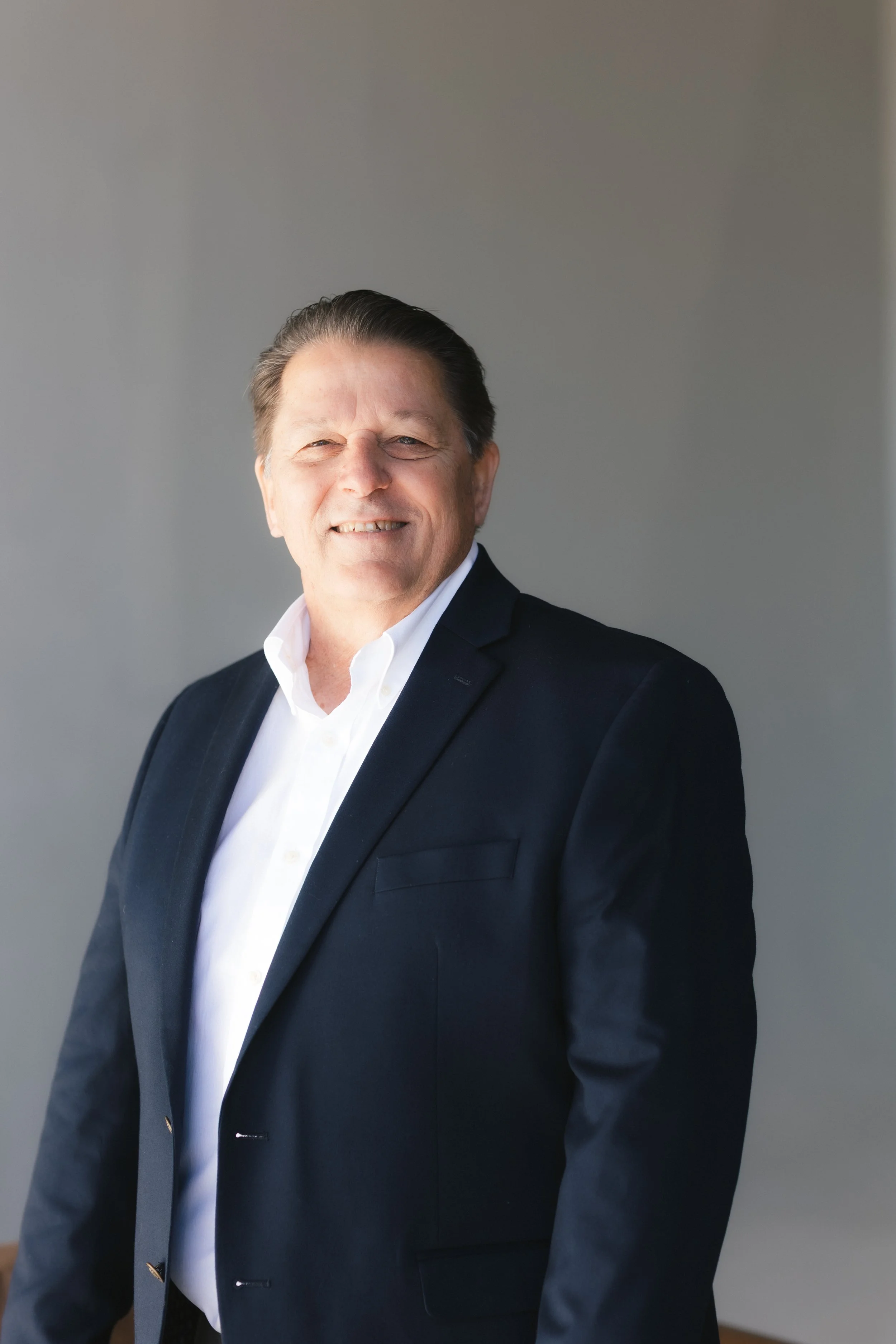 A smiling middle-aged man in a black suit jacket and white dress shirt standing against a plain wall.