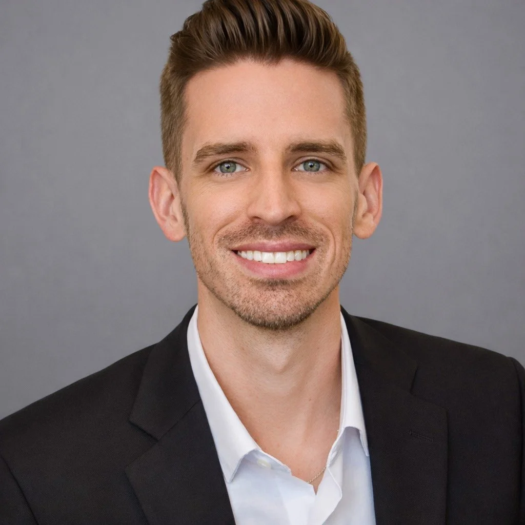 A young man in a black suit and white shirt smiling against a grey background.