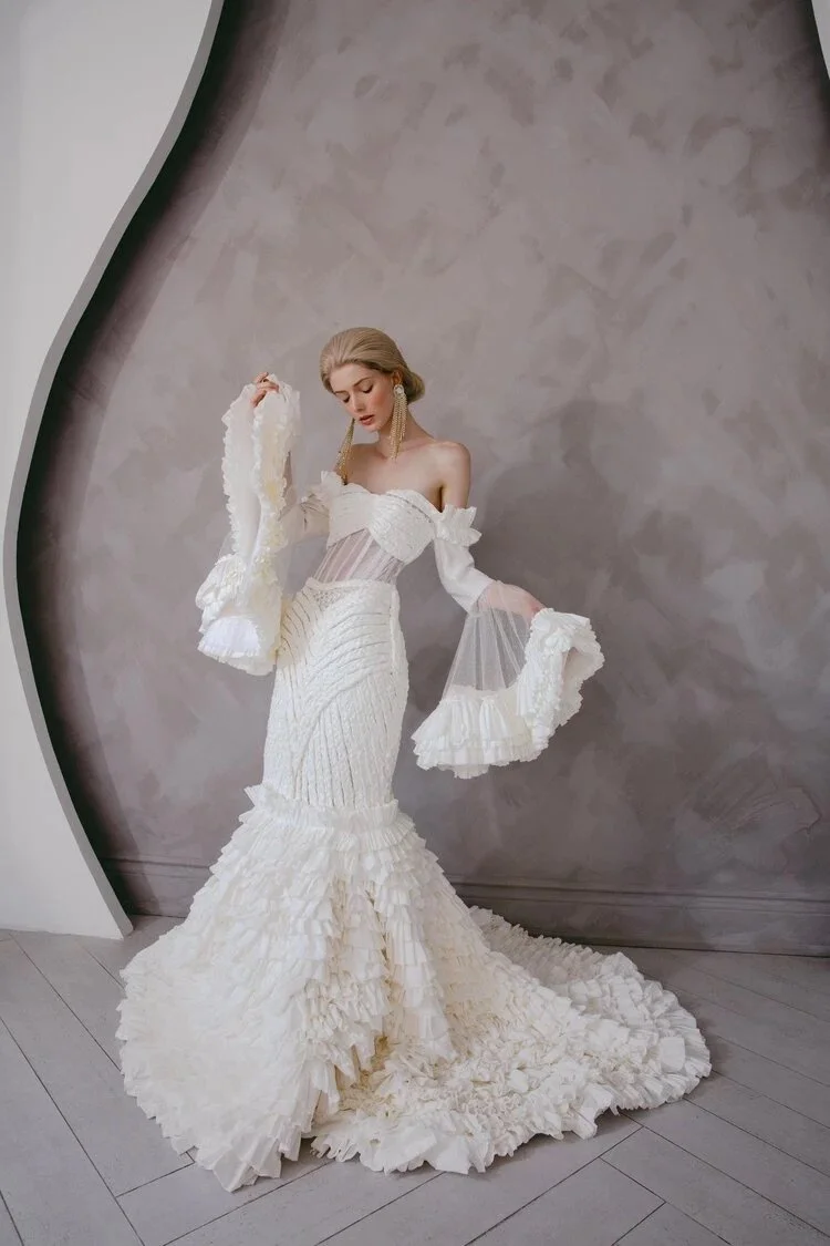 Model in intricate white ruffled gown standing against textured wall.