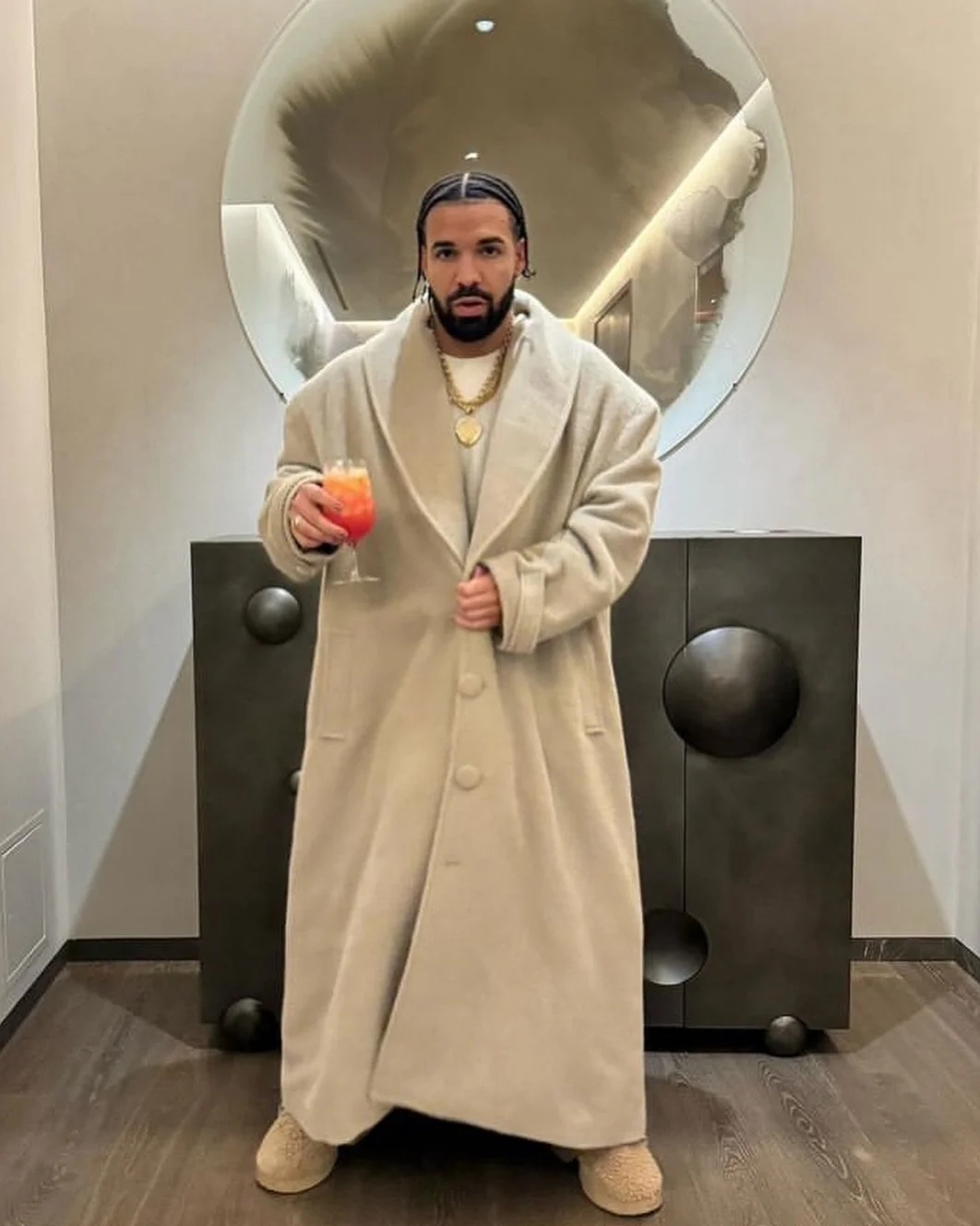 A man wearing a long beige coat stands in a modern room, holding a glass of orange beverage. Behind him, there is a round mirror and a black cabinet with circular patterns.