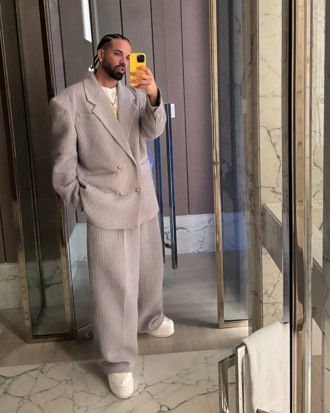 Person taking a mirror selfie wearing a light gray oversized suit and white sneakers, holding a yellow phone, standing in a modern bathroom with marble and glass decor.